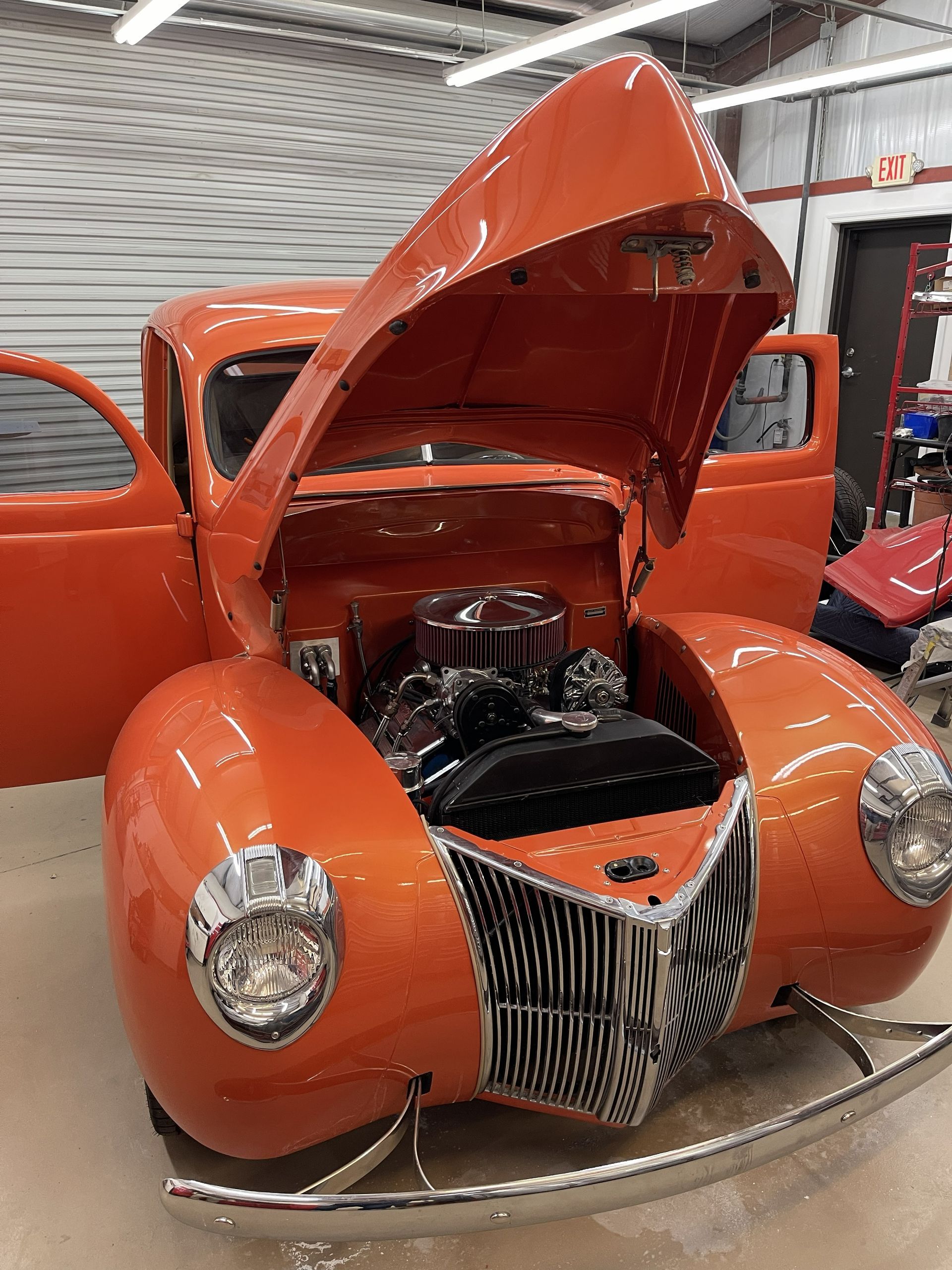 Bright orange vintage hot rod with an open hood, chrome grille, and headlights, parked inside a garage.