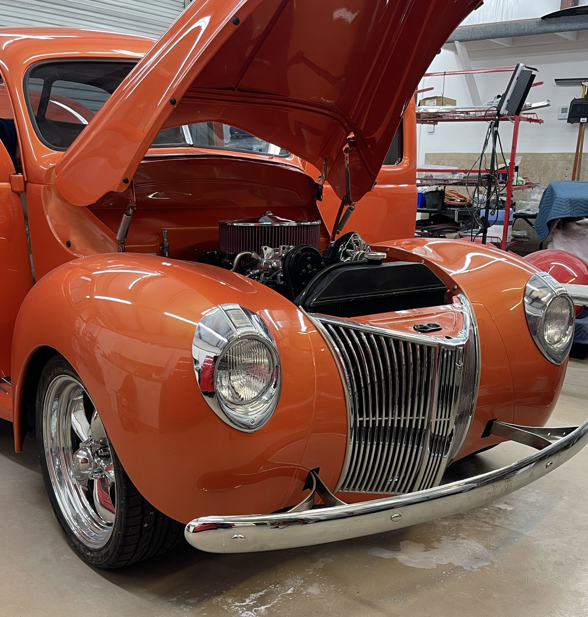 A bright orange vintage Ford coupe with a chrome grille and open hood sits in a workshop.