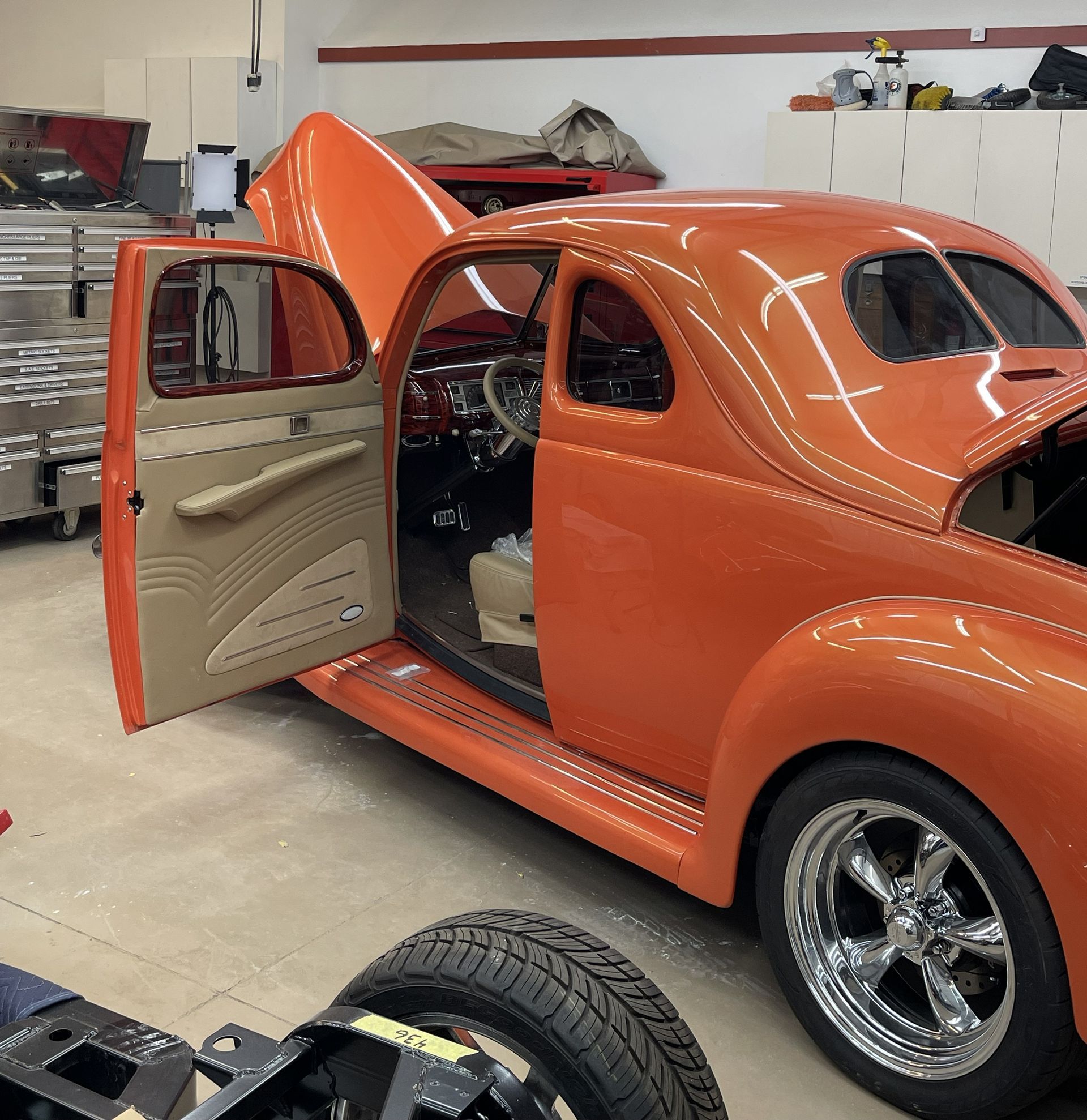 A glossy orange vintage hot rod car parked inside a garage with its hood and driver-side door open.