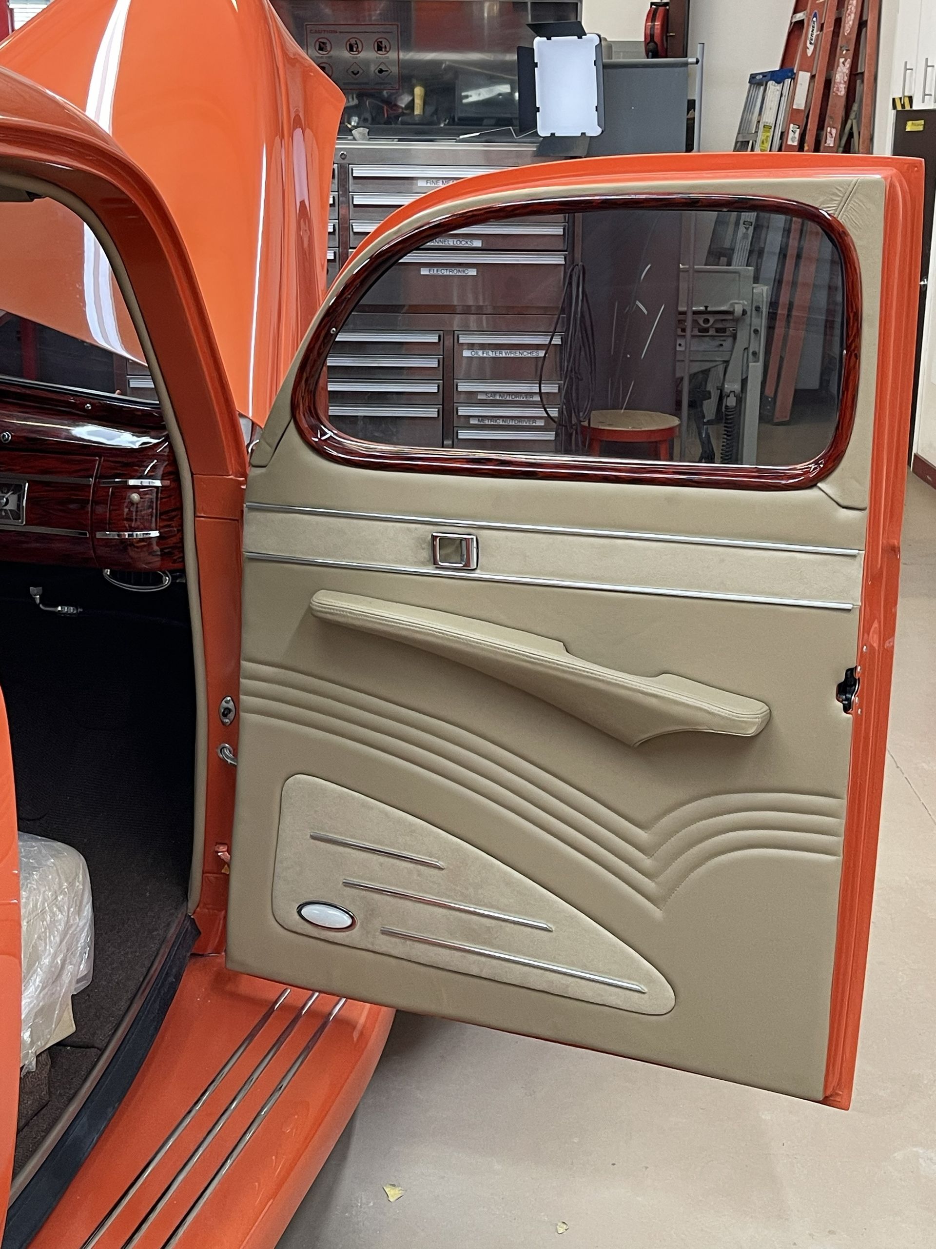 Open orange car door with custom tan upholstered interior, including a horizontal armrest and stitched panel designs.