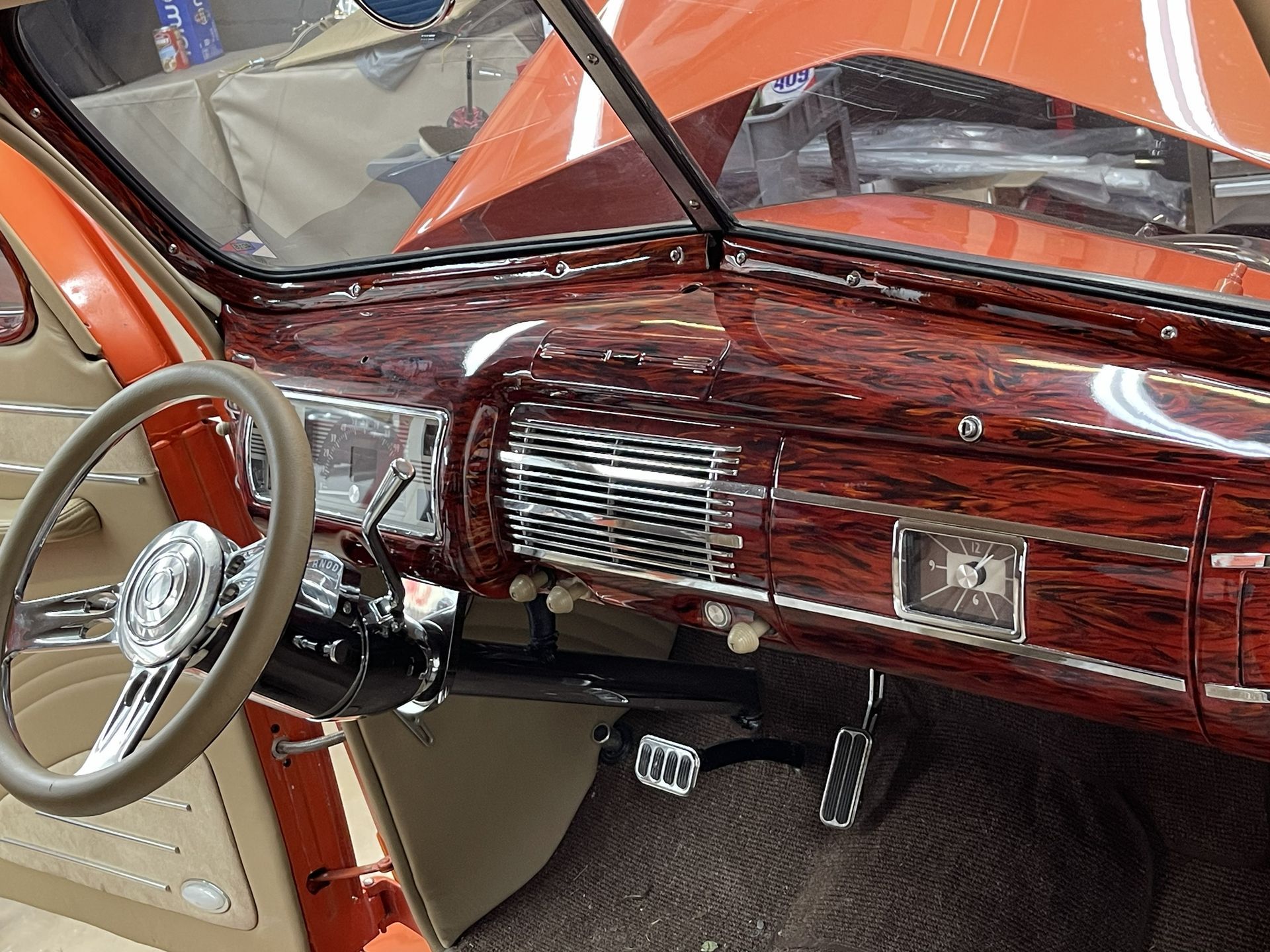 Interior view of a vintage car featuring a polished wood-grain dashboard, tan steering wheel, and pedals.