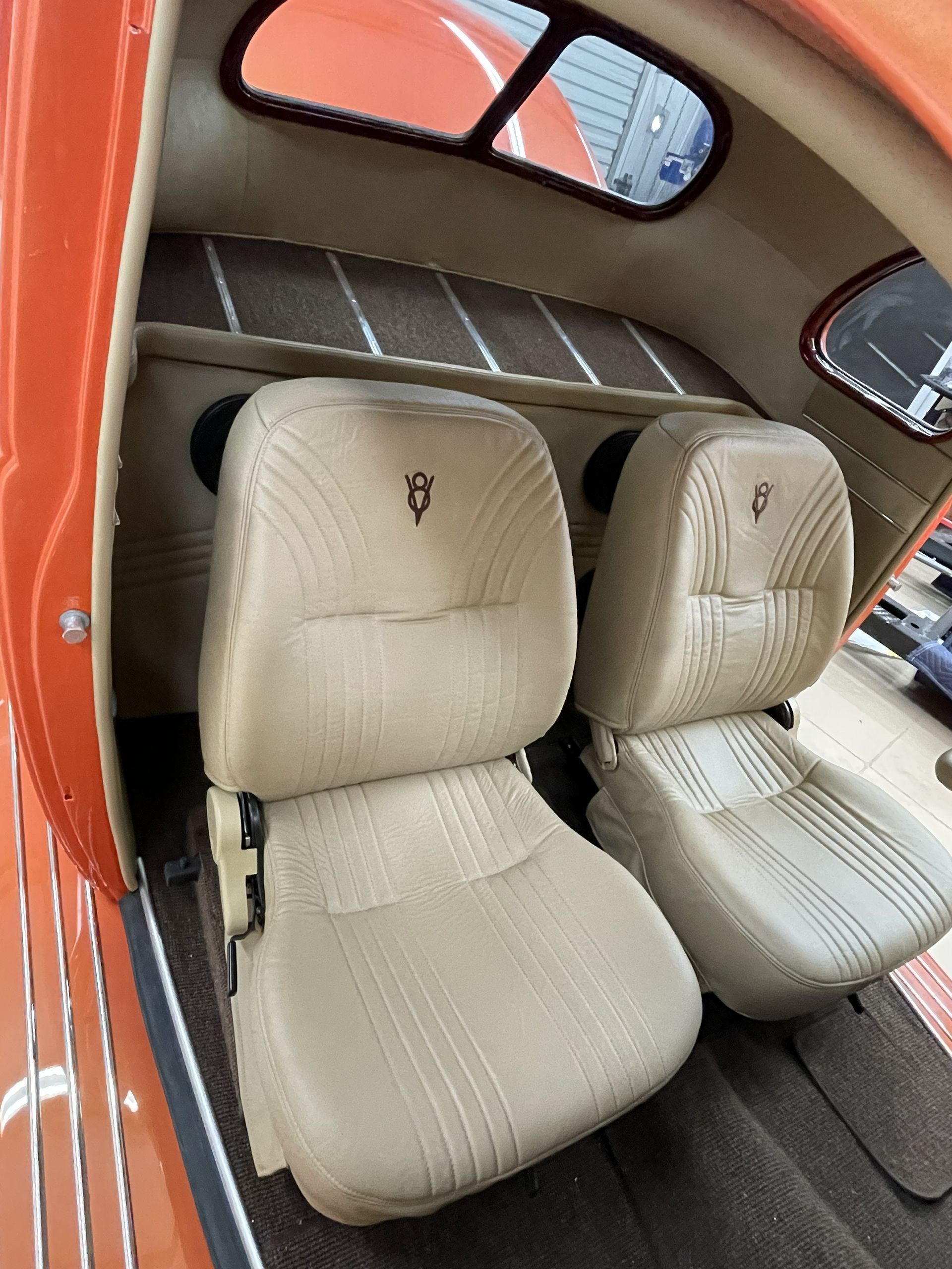 Tan leather bucket seats with a gold emblem inside the orange interior of a classic car with a dark carpet floor.