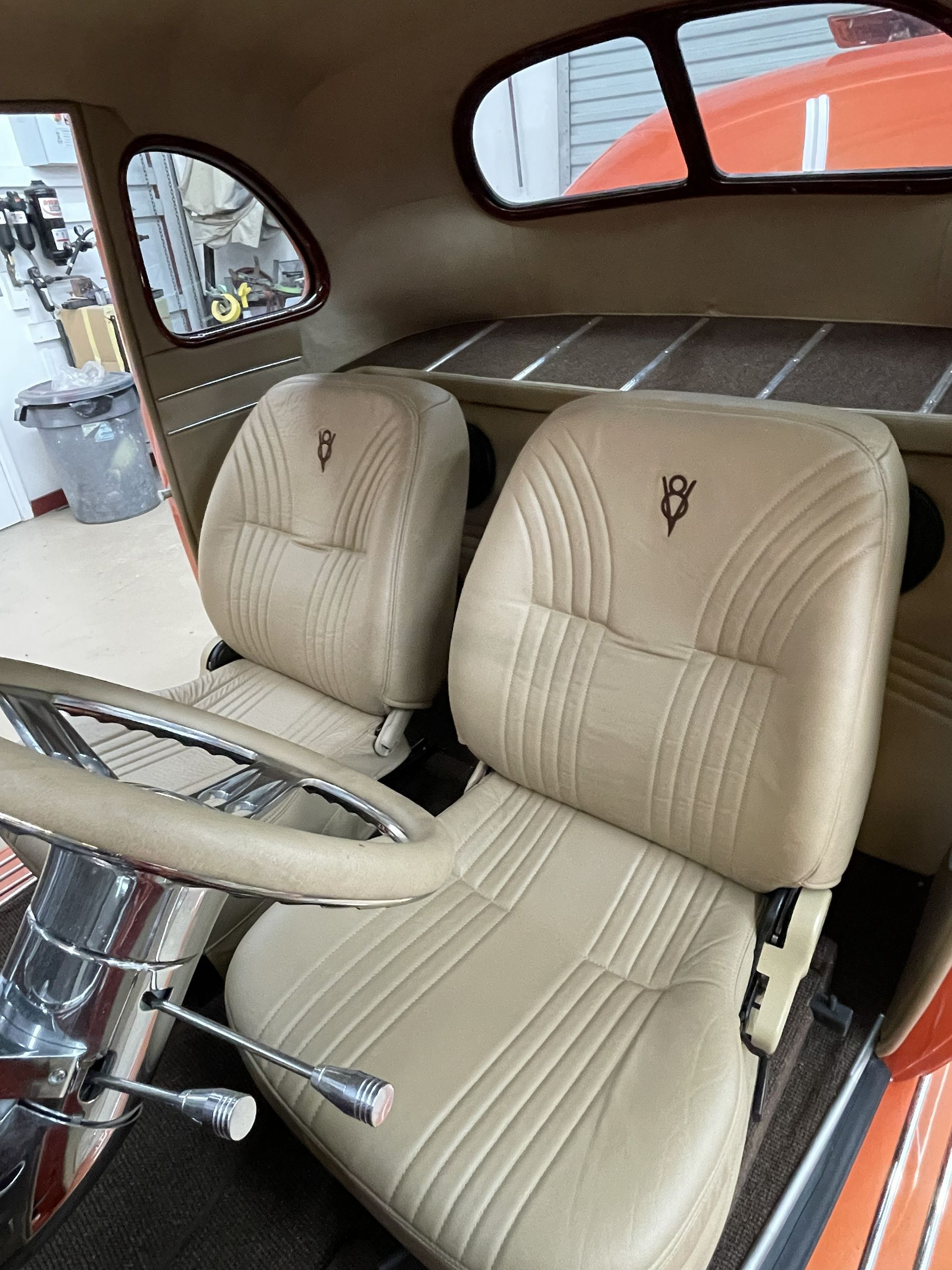 Interior of a classic car featuring beige leather bucket seats with a V8 emblem, a steering wheel, and a rear parcel shelf.