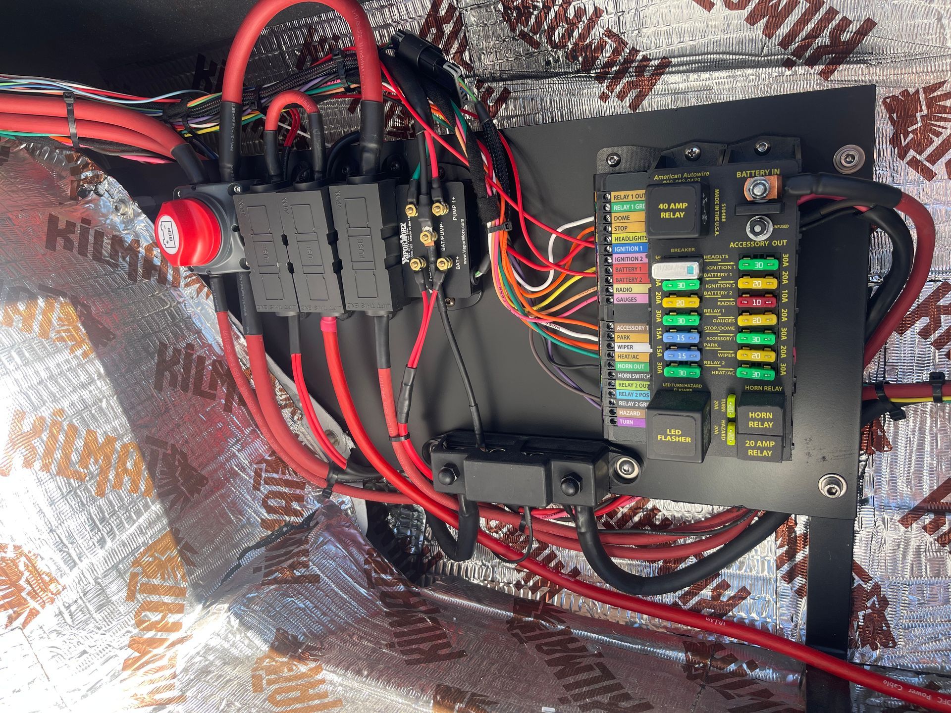 A custom automotive electrical panel mounted on a sound-dampened wall, featuring a fuse block, relays, and red wiring.