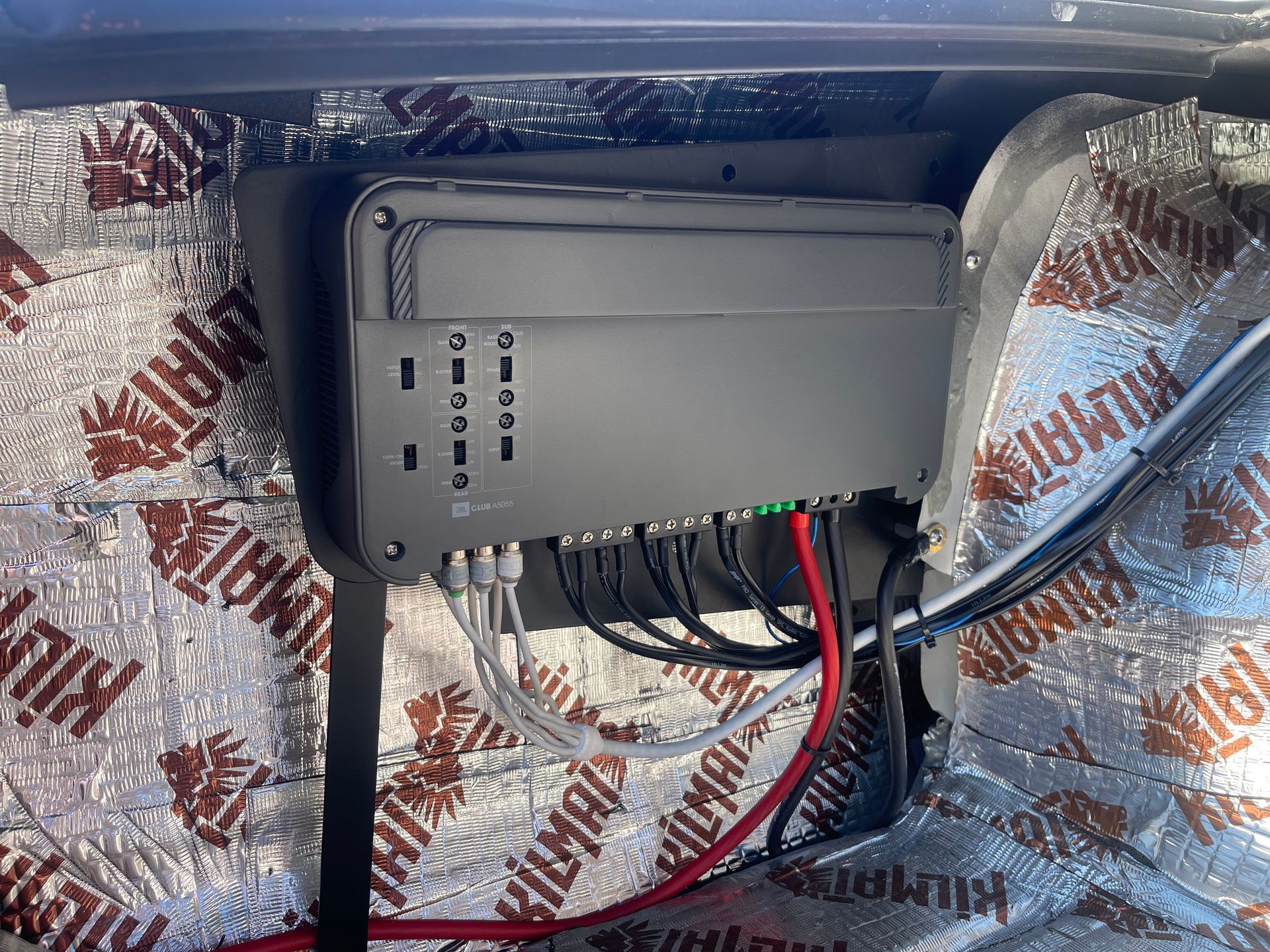 A black car audio amplifier mounted on a vehicle wall covered in silver sound-deadening material with attached wiring.