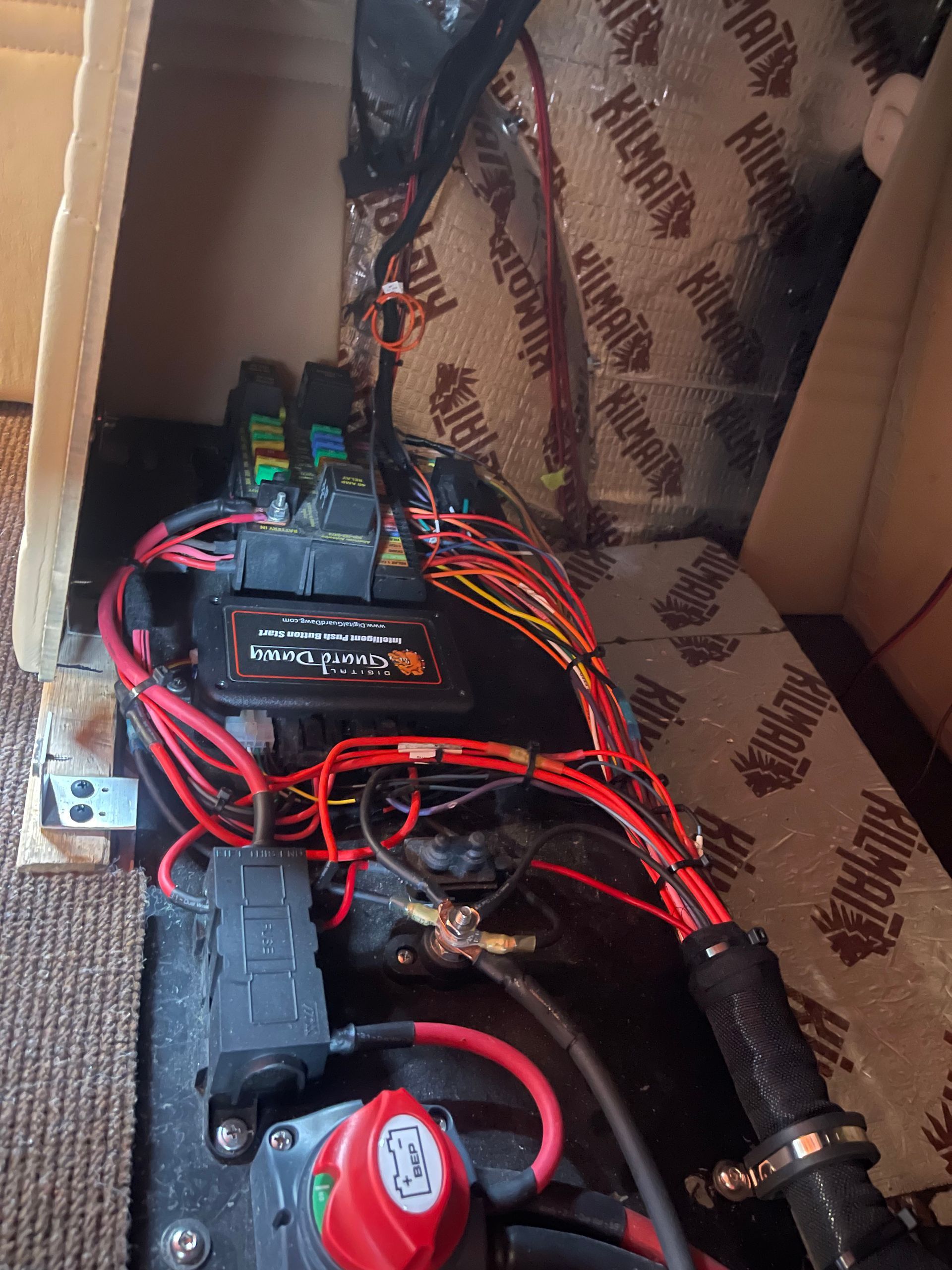 A close-up of a complex vehicle electrical system, featuring wires, a fuse box, a red battery switch, and Kilmat insulation.