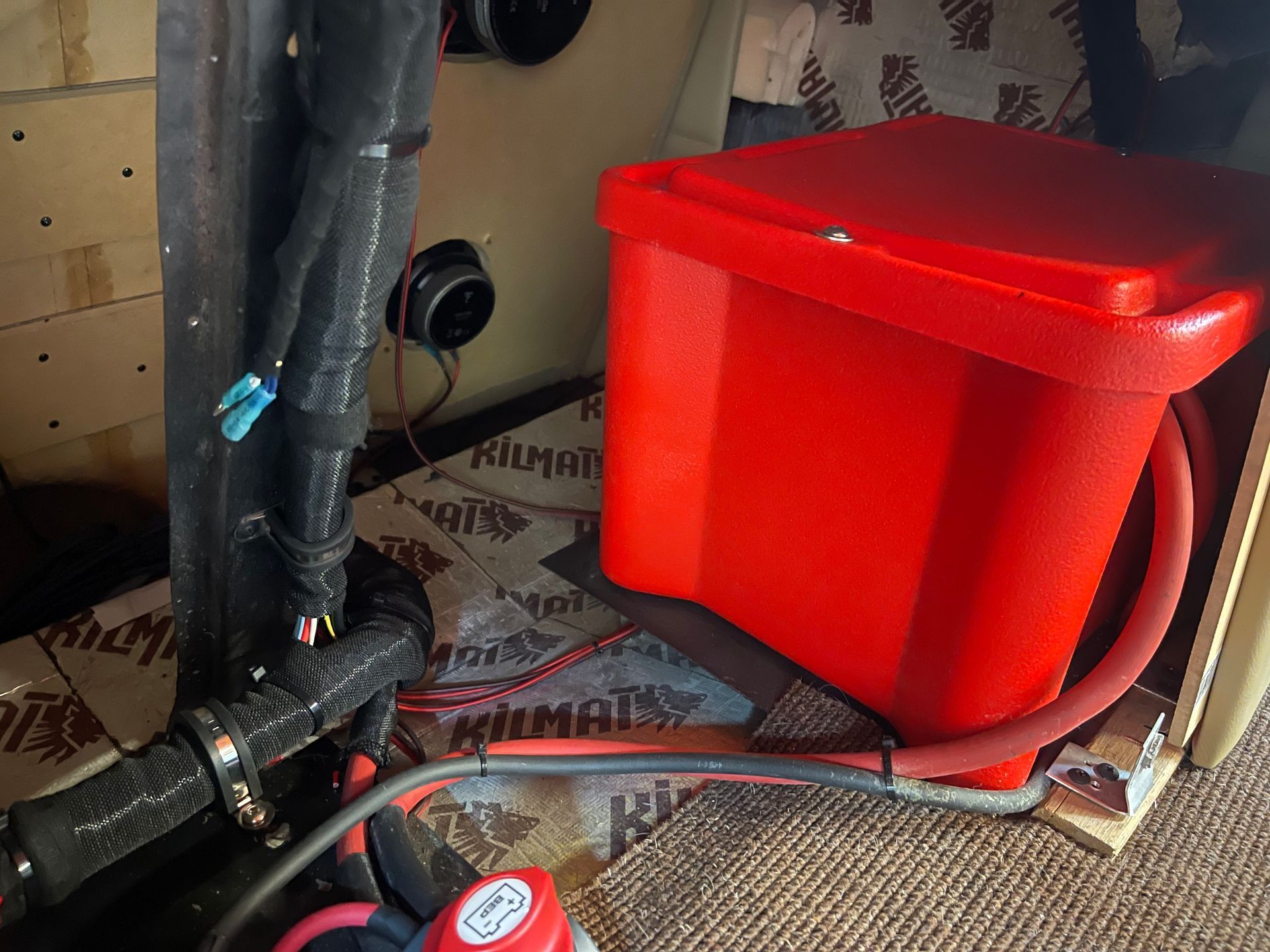 A bright red plastic battery box sits on a floor lined with sound-deadening material, alongside cables and vehicle wiring.