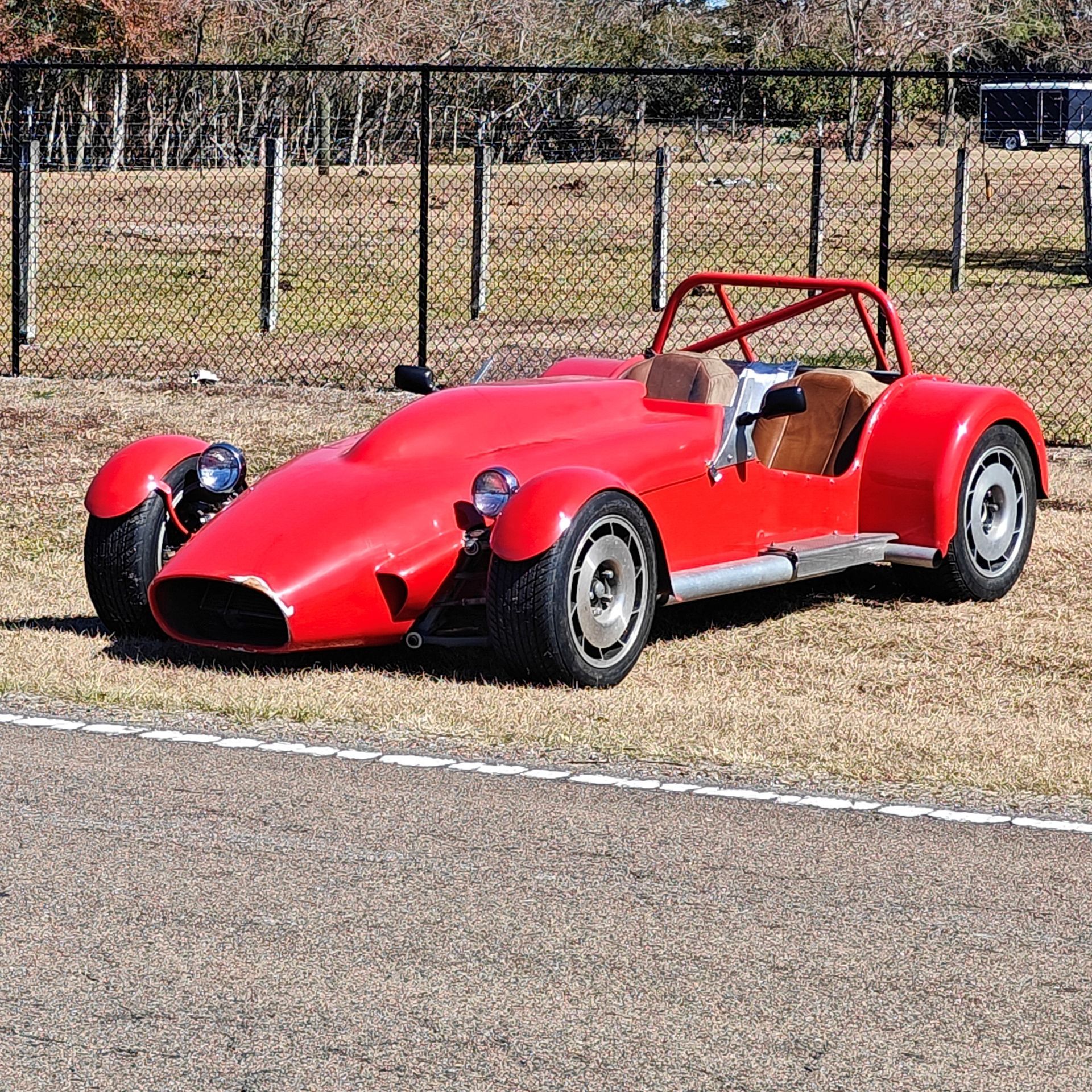 A bright red open-cockpit sports car parked on grass beside an asphalt track with a wire fence in the background.