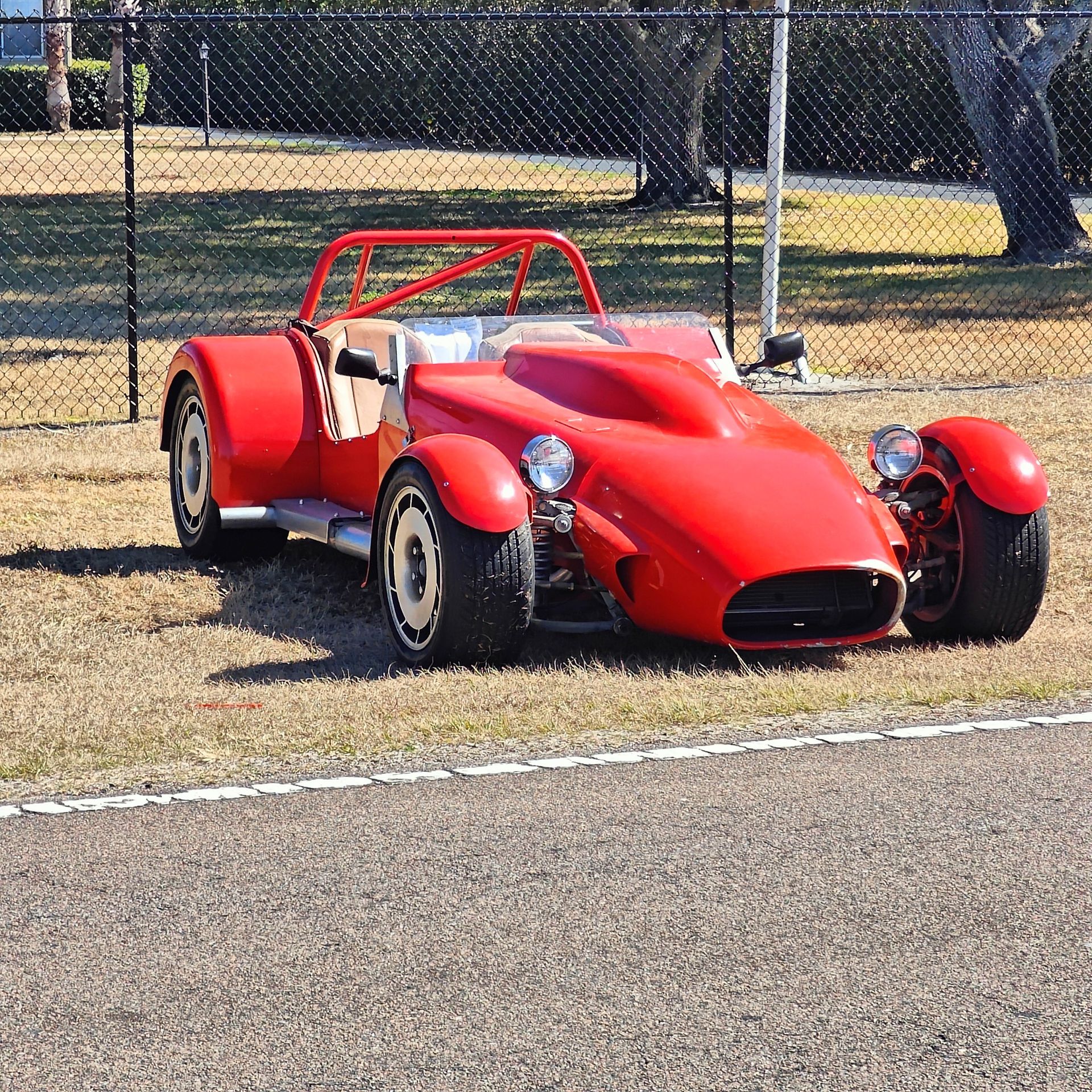 A bright red, open-cockpit sports car parked on grass next to a chain-link fence on a sunny day.