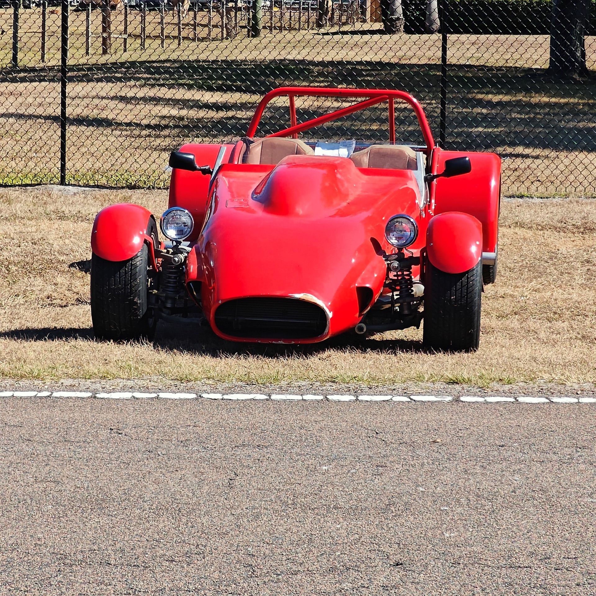 A bright red, open-cockpit sports car with a roll cage parked on an asphalt track in front of a chain-link fence.