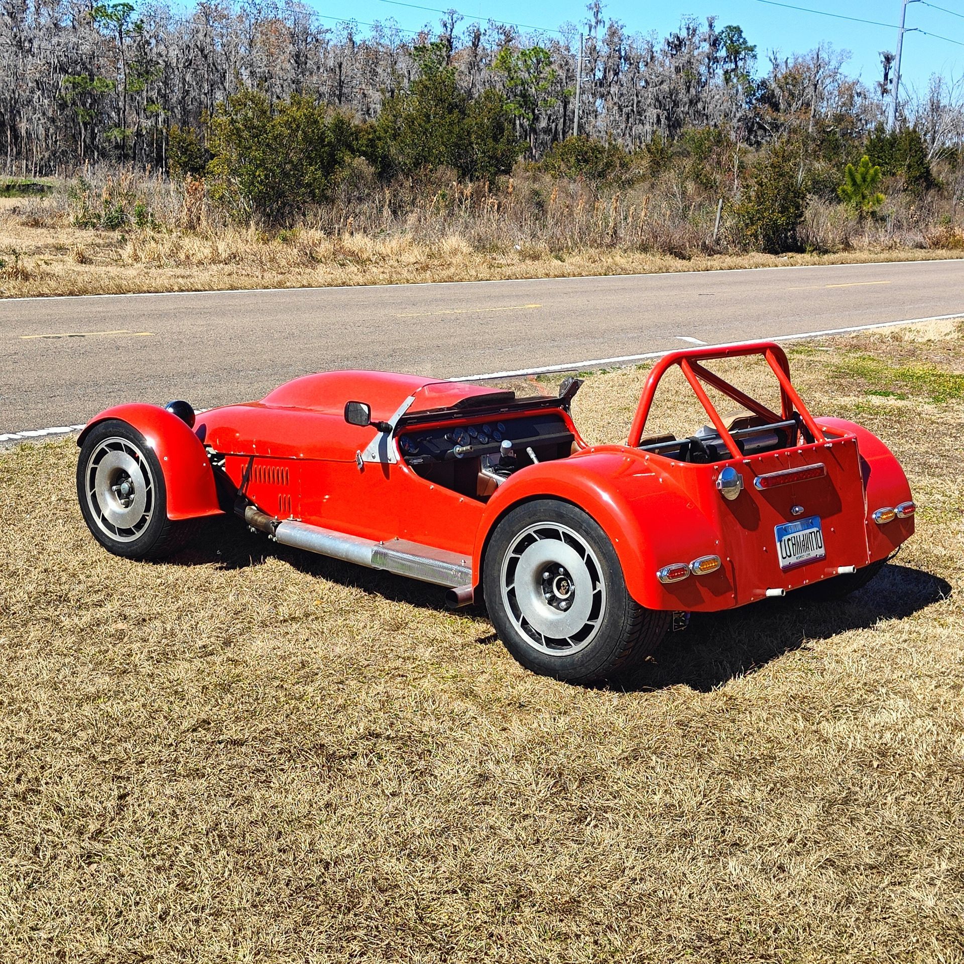 A bright red, two-seater roadster with a roll bar parked on grass next to a road under a clear blue sky.