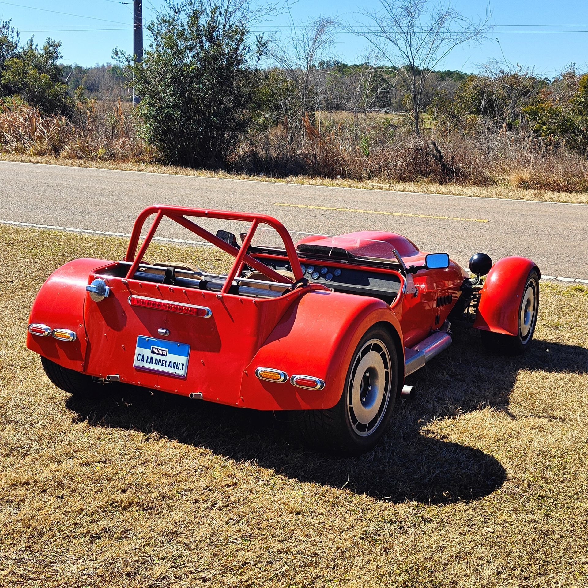 A bright red vintage-style sports car parked on a grass verge beside a road under a clear blue sky.