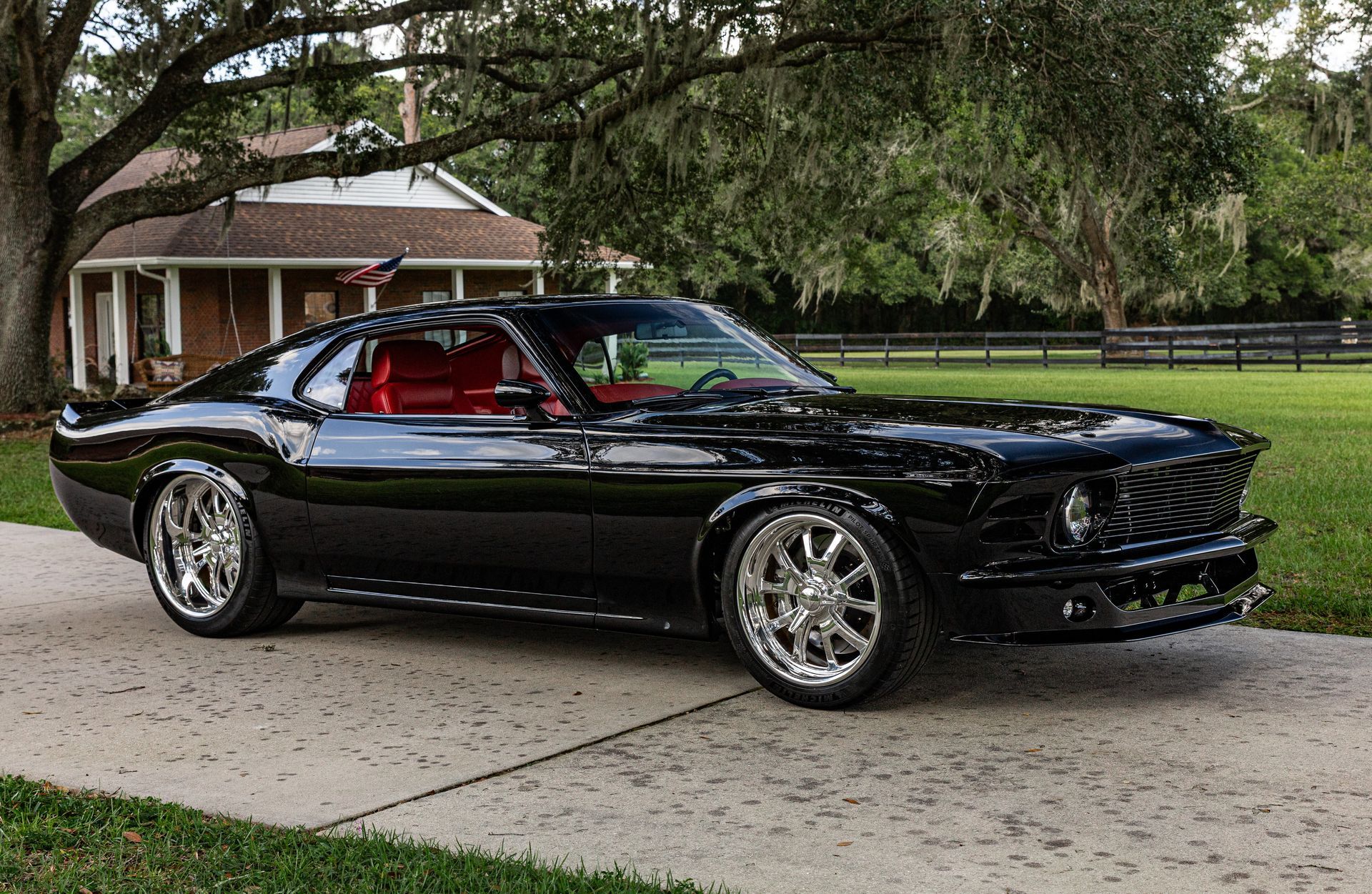 A sleek, black custom Ford Mustang with chrome wheels parked on a driveway in front of a house and large trees.