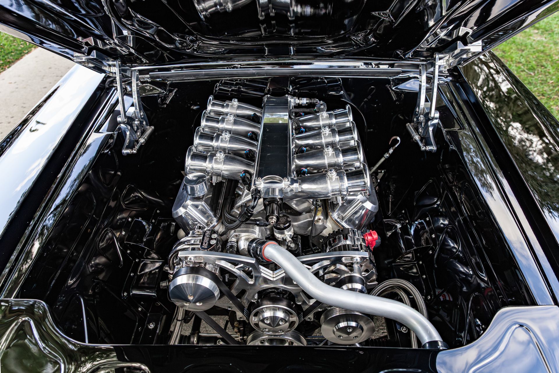 A high-angle view of a shiny, black classic car engine bay featuring a polished chrome V8 engine with intake stacks.