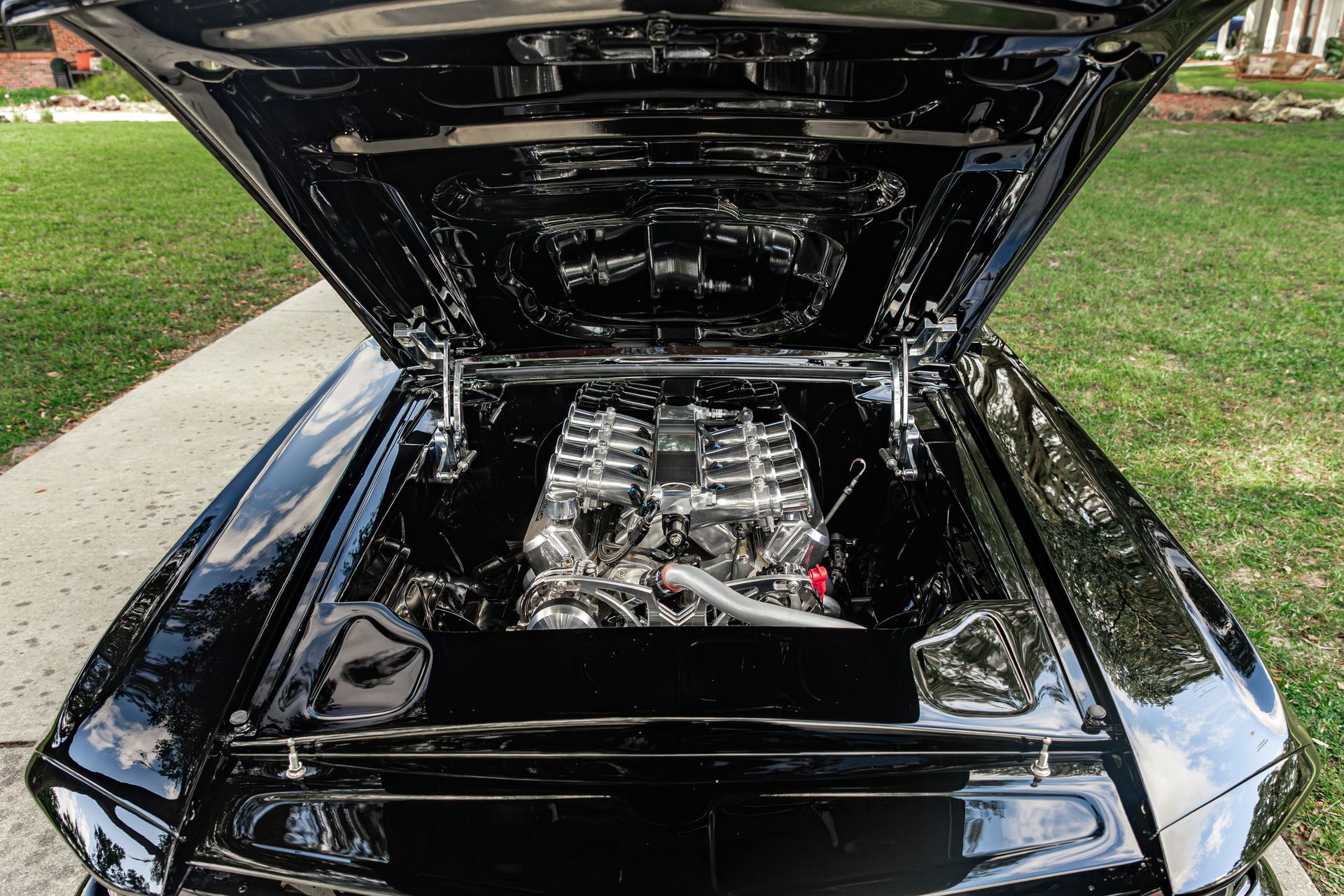 A close-up view of the polished black engine bay of a classic car, with its hood propped open outdoors.