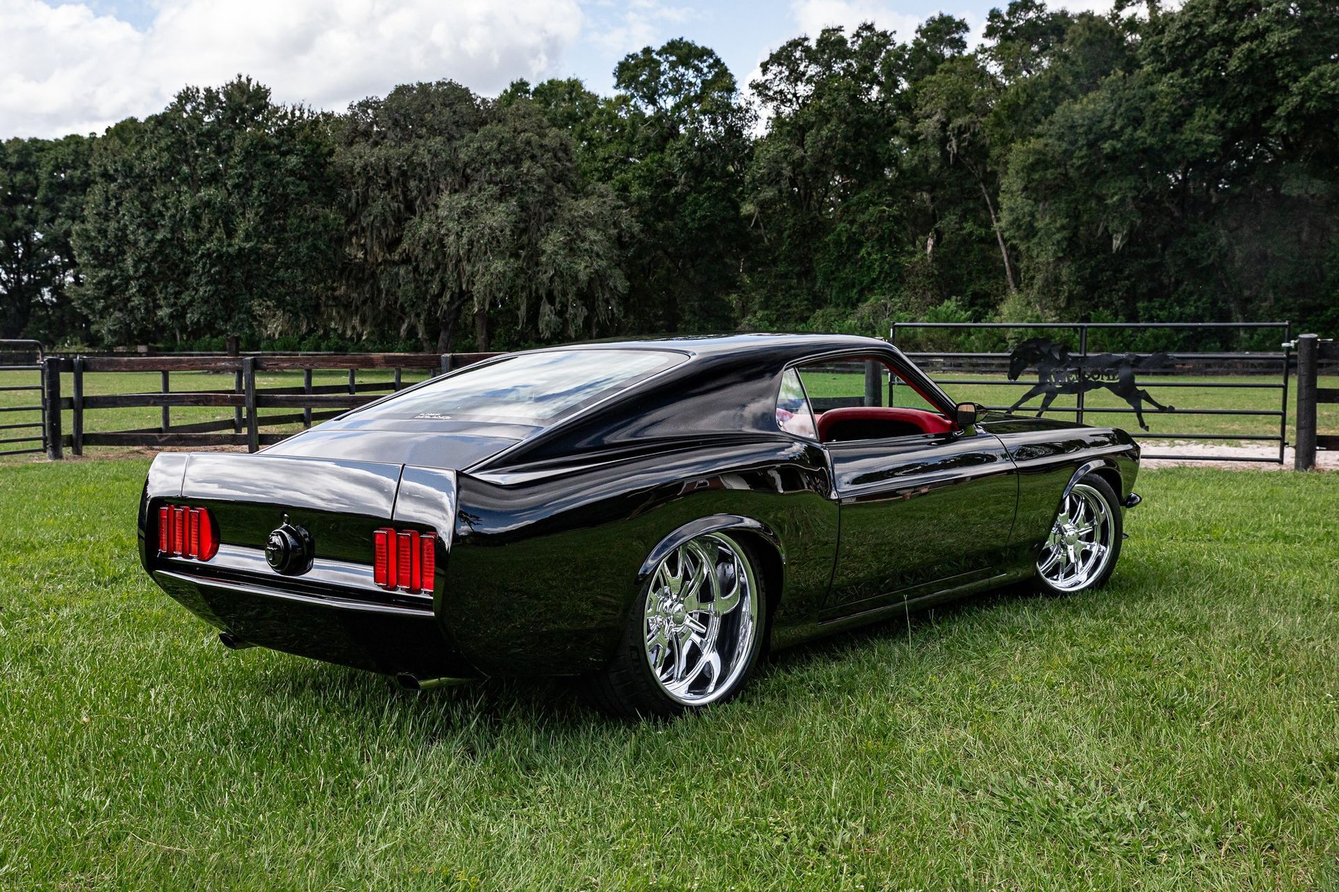 A glossy black vintage Mustang muscle car parked on a grassy field in front of a wooden fence.
