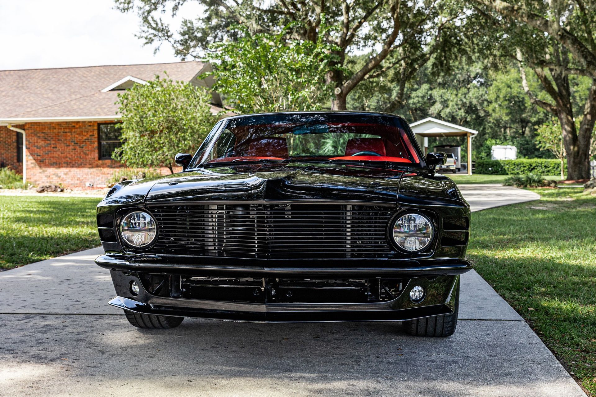A black classic muscle car parked on a paved driveway in front of a brick house on a sunny day.