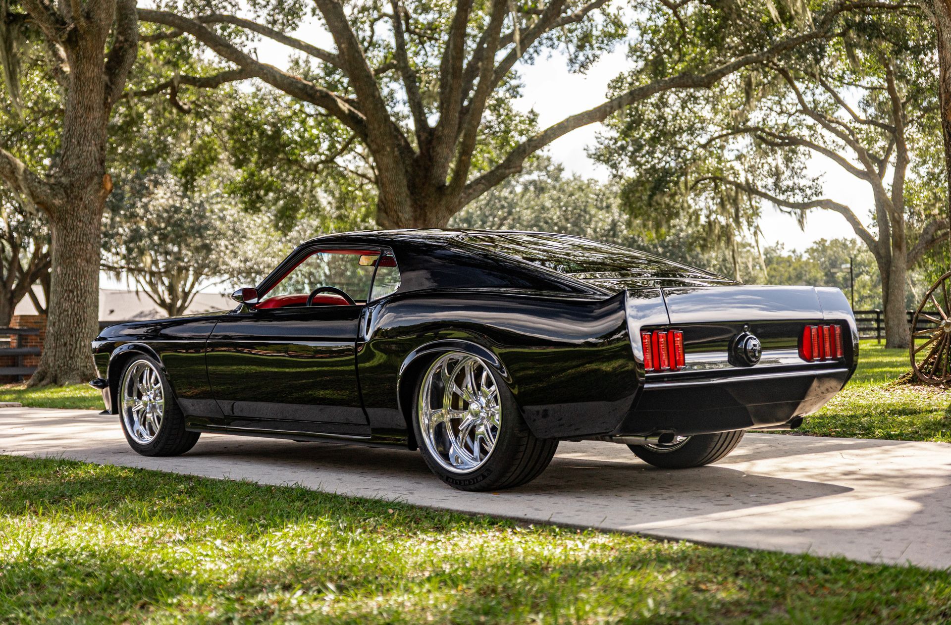 A black custom 1969 Ford Mustang with chrome wheels parked on a gravel path under mossy oak trees.