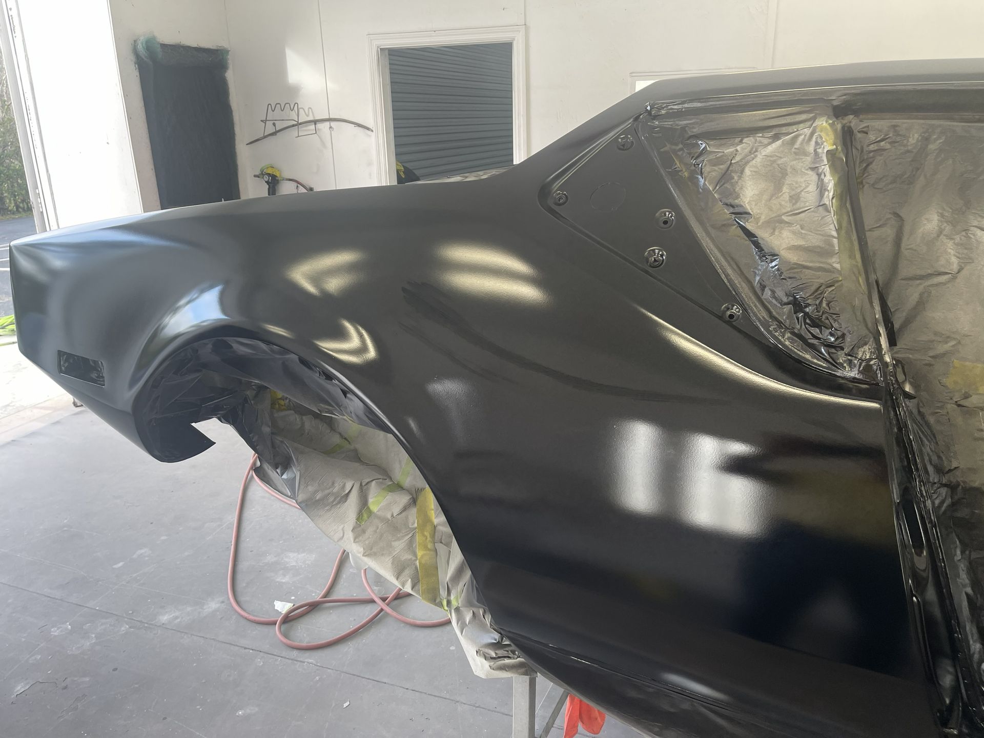 A black car body panel being painted in a shop, shown with masked wheel arches and reflective paint finish.