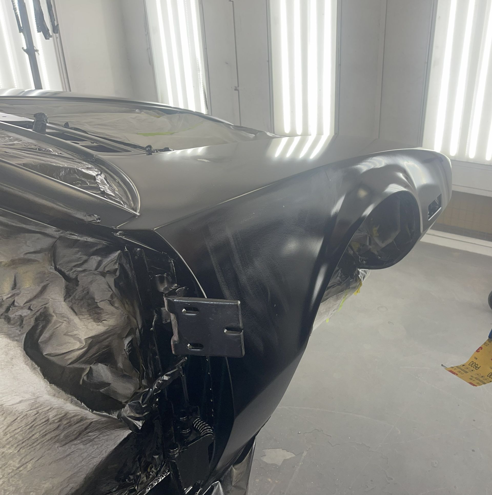The front fender and hood of a black car are shown in a bright, indoor workshop, appearing freshly painted or primered.