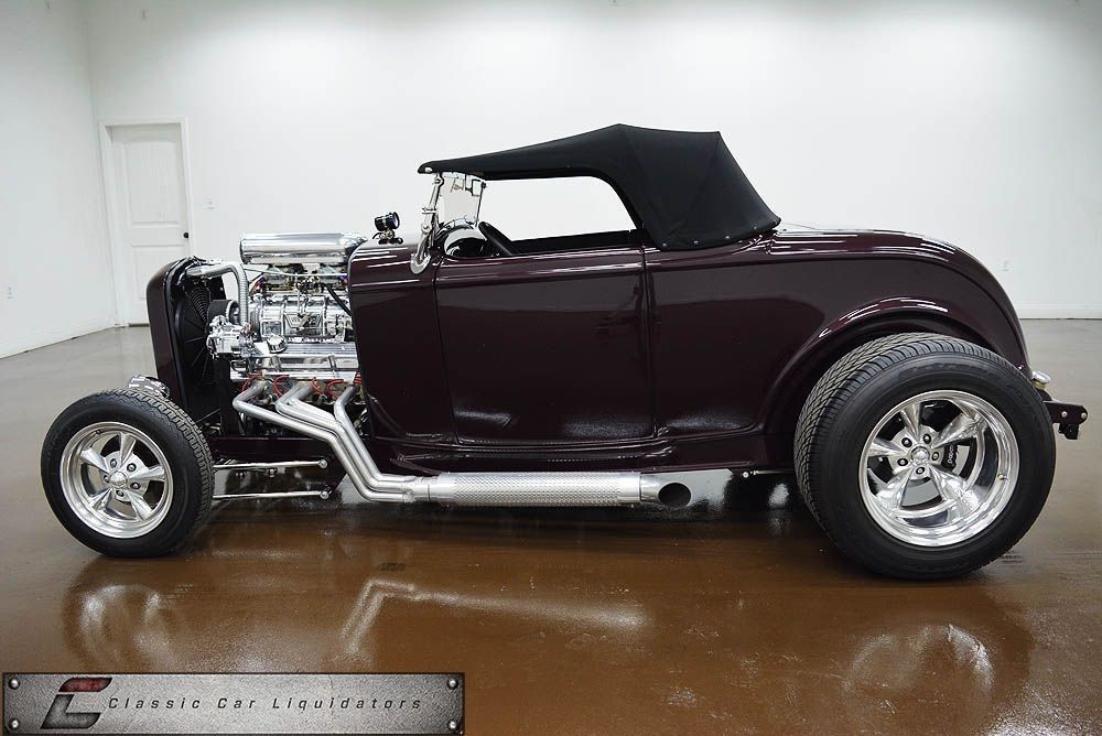 A deep maroon vintage roadster hot rod with a black soft top and chrome engine sits in a showroom.