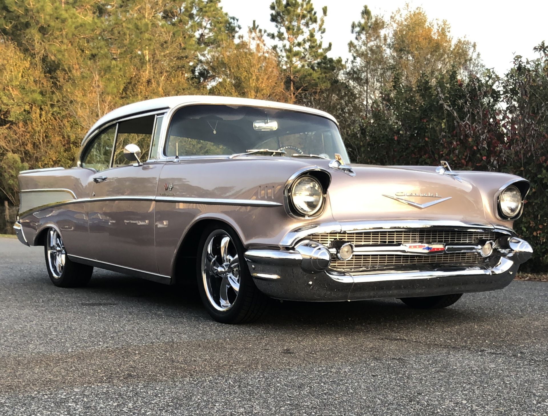 A classic, two-tone mauve and white 1957 Chevrolet Bel Air parked on asphalt with trees in the background.