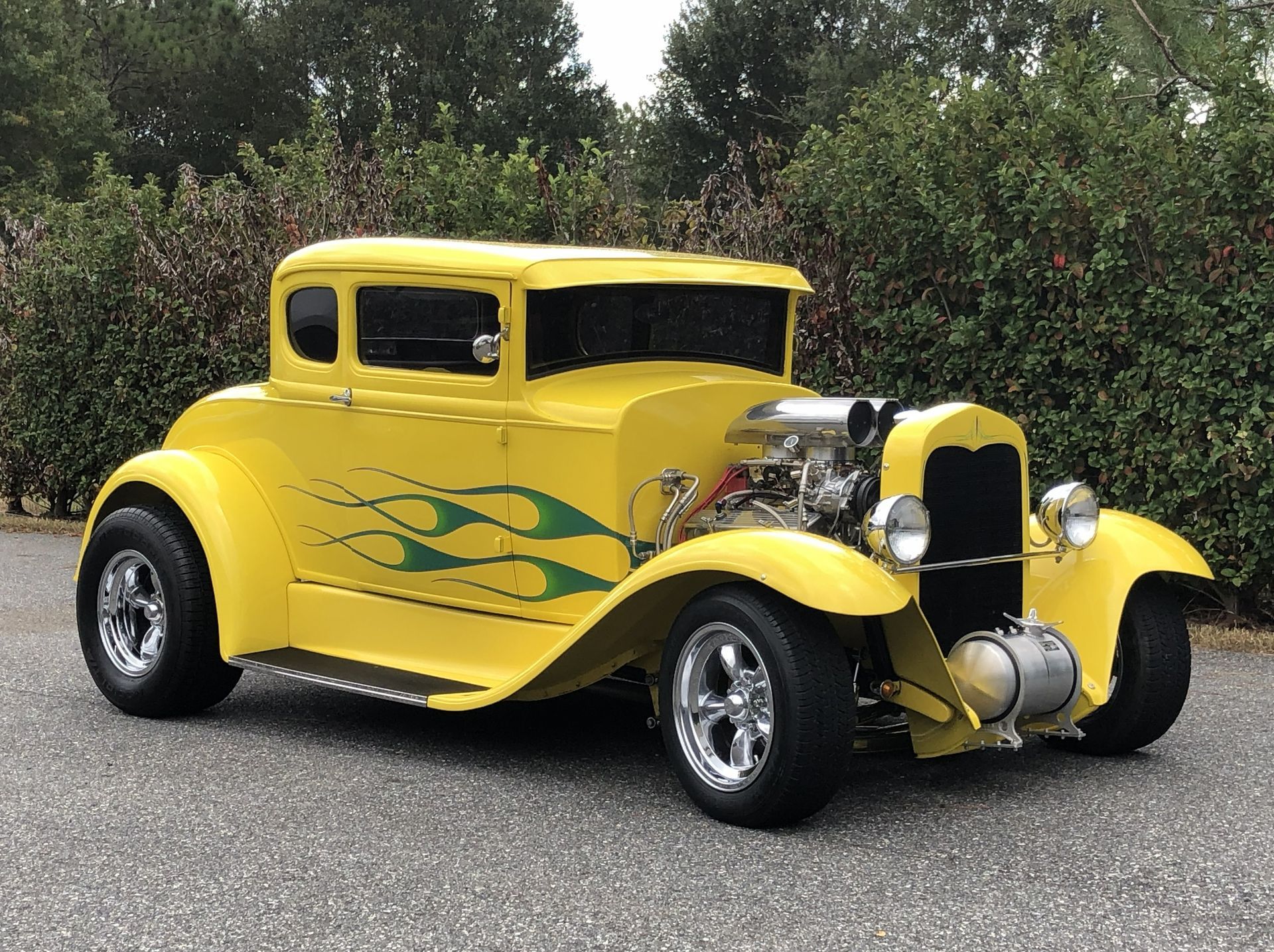 A bright yellow hot rod coupe with green flame decals, a large chrome engine, and chrome wheels parked on gravel.