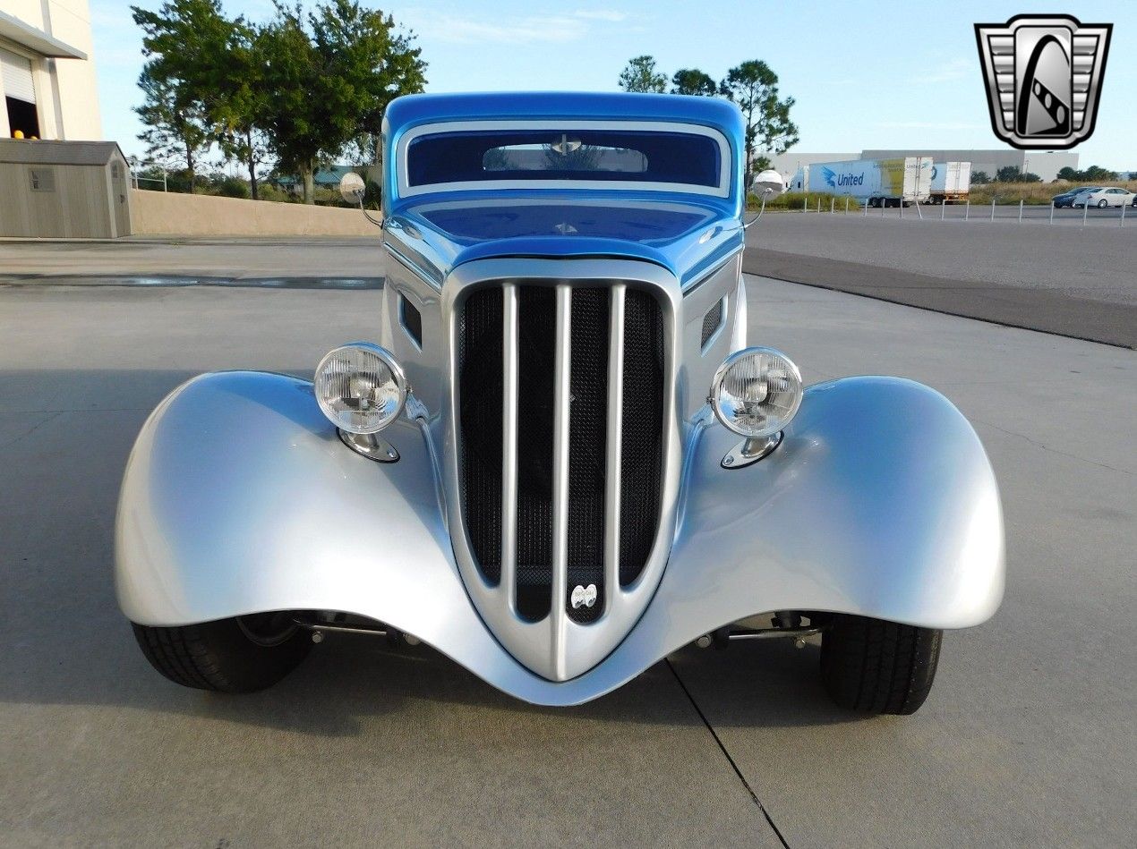 Front view of a custom silver and blue hot rod car parked on an asphalt lot outdoors.