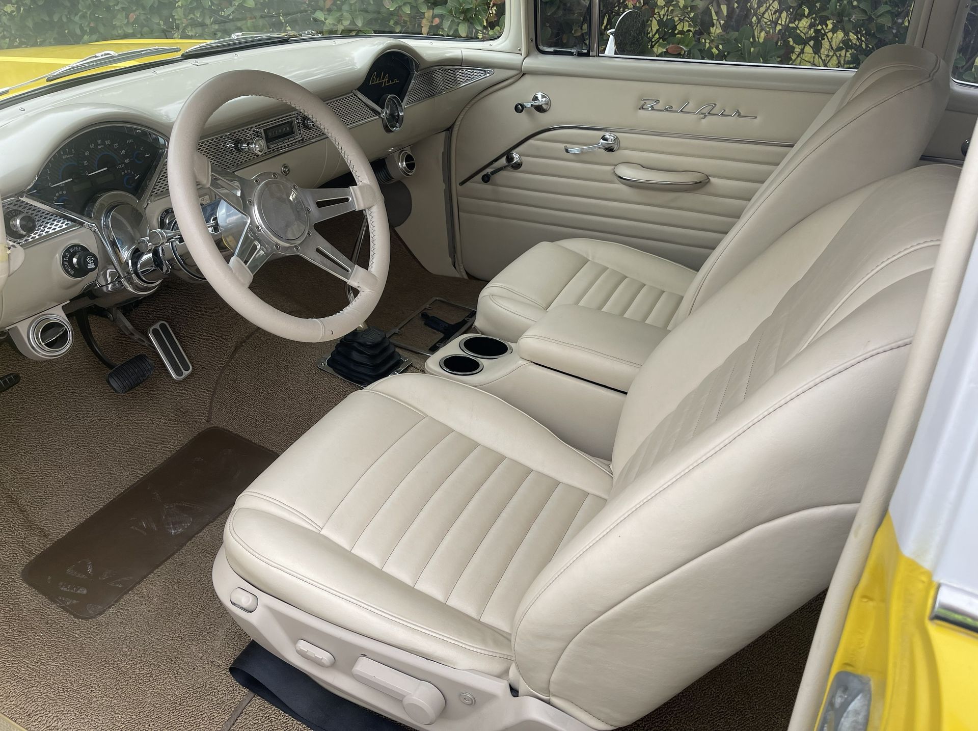 The cream-colored, leather interior of a classic car, featuring a white steering wheel and textured floor mats.