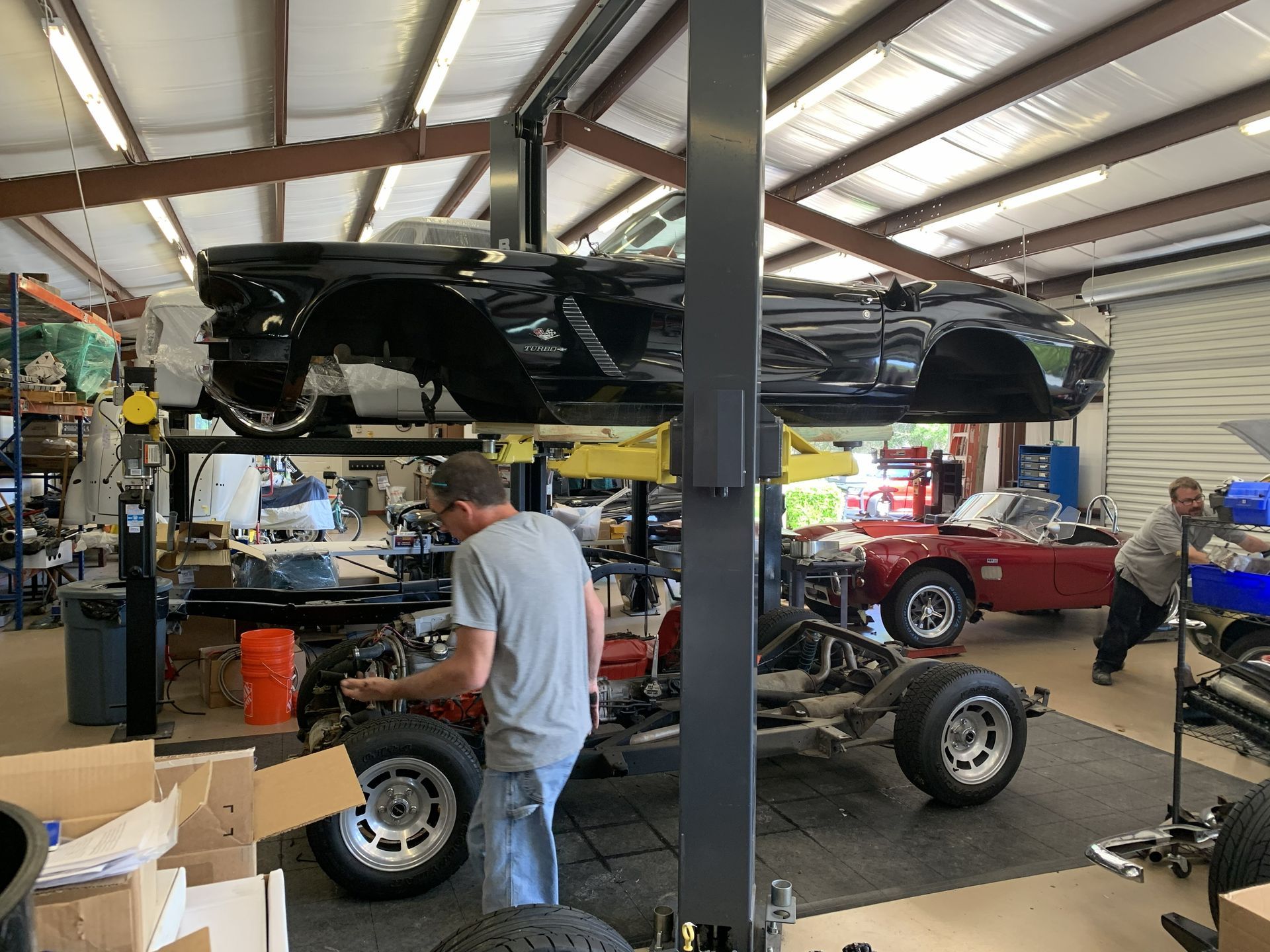Mechanics work on a black vintage car body elevated on a lift in a workshop, with a red convertible parked nearby.