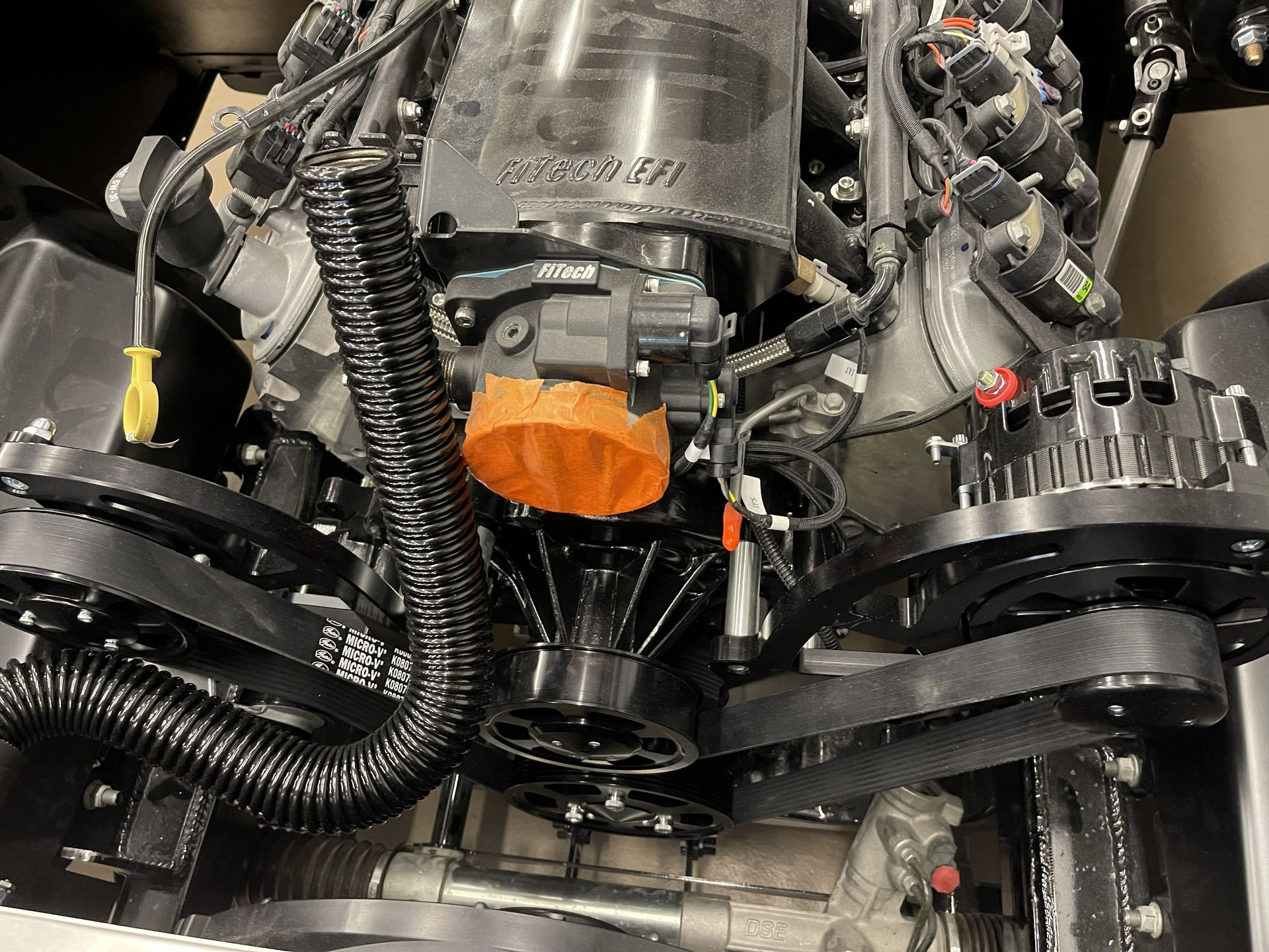 Close-up of a high-performance black engine inside a vehicle, featuring belts, pulleys, and an orange intake cap.