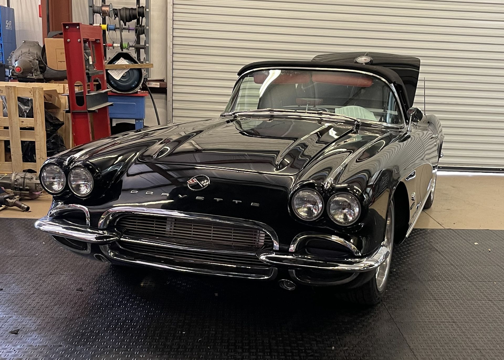 A black vintage Chevrolet Corvette convertible parked inside a garage on a black diamond-plate floor.