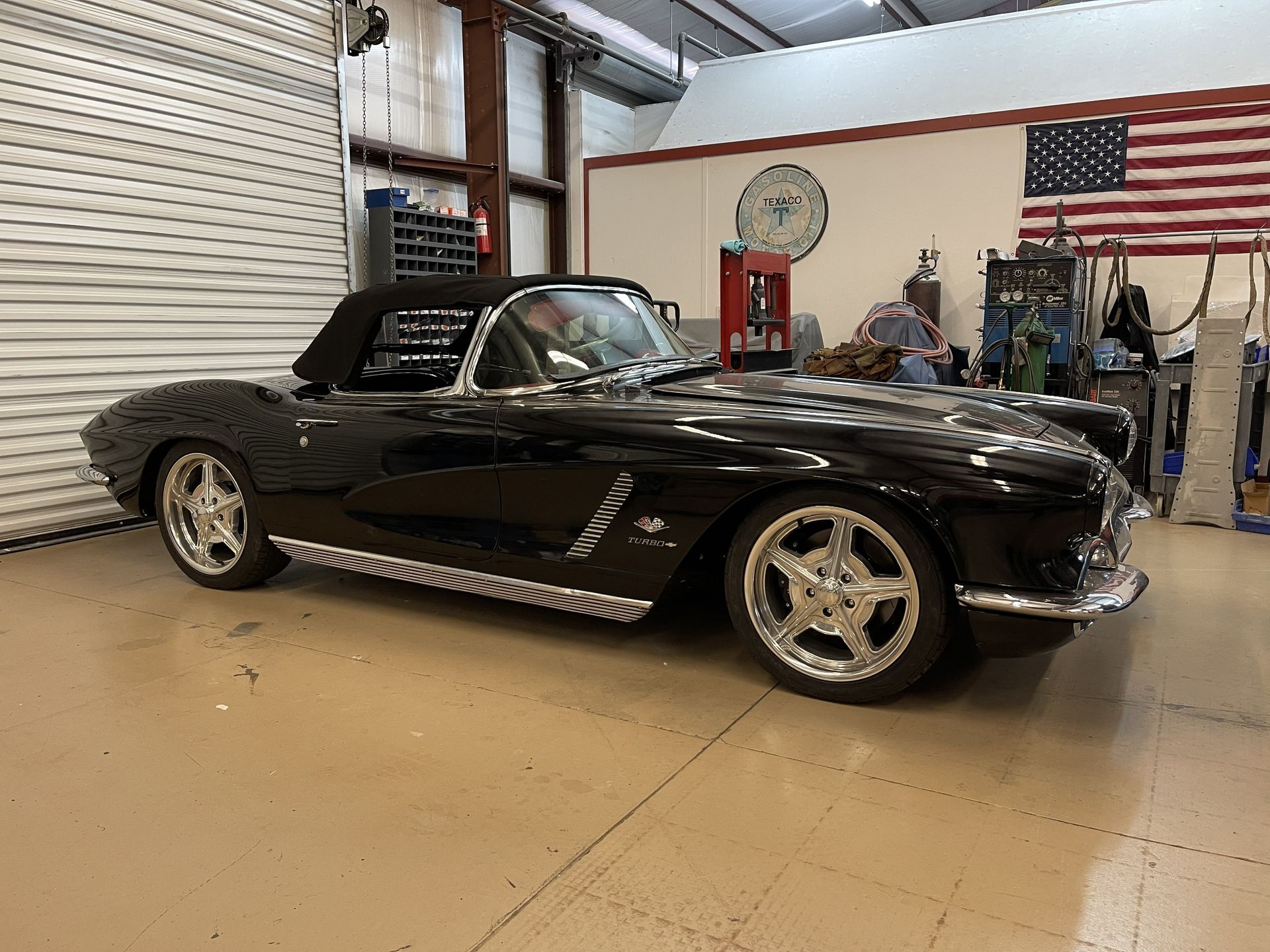 A shiny black vintage convertible Corvette parked in a garage with a US flag on the wall.
