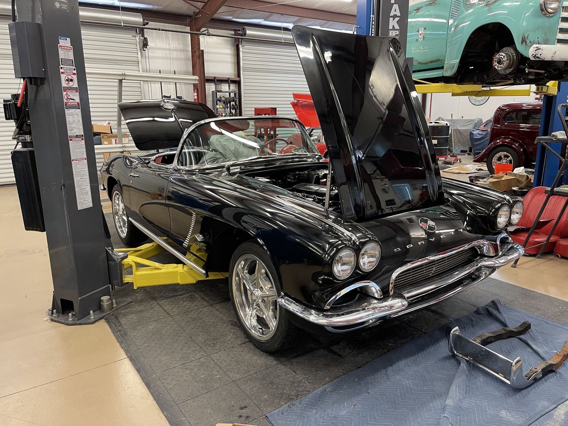 A black classic Corvette with its hood raised on a yellow hydraulic automotive lift inside a repair shop.