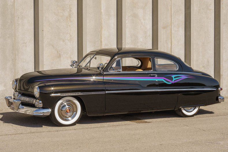 A vintage black custom Mercury coupe with whitewall tires and teal pinstriping parked against a ribbed concrete wall.
