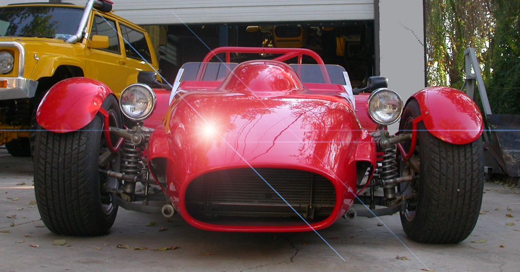A bright red kit car parked on a concrete driveway in front of a garage, with a yellow off-road vehicle partially visible.