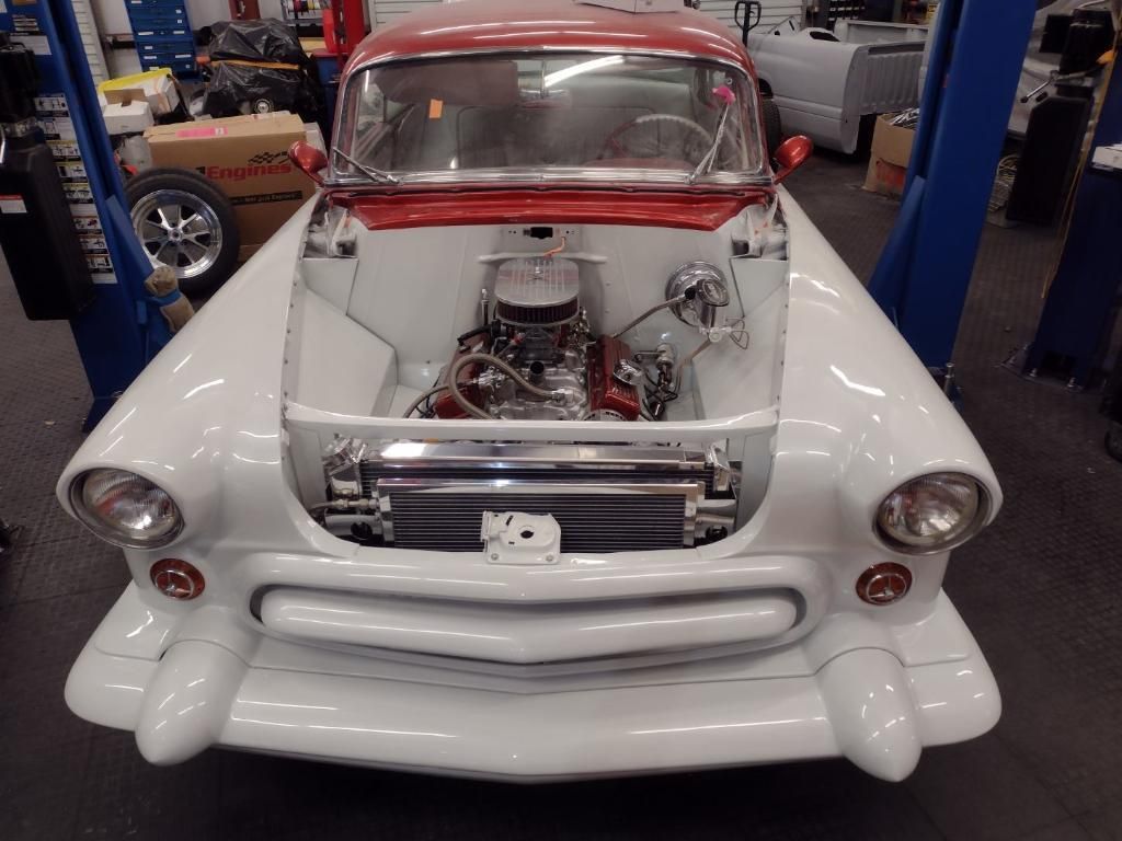 A classic car with its hood removed, revealing a detailed engine inside a custom white engine bay in a garage.