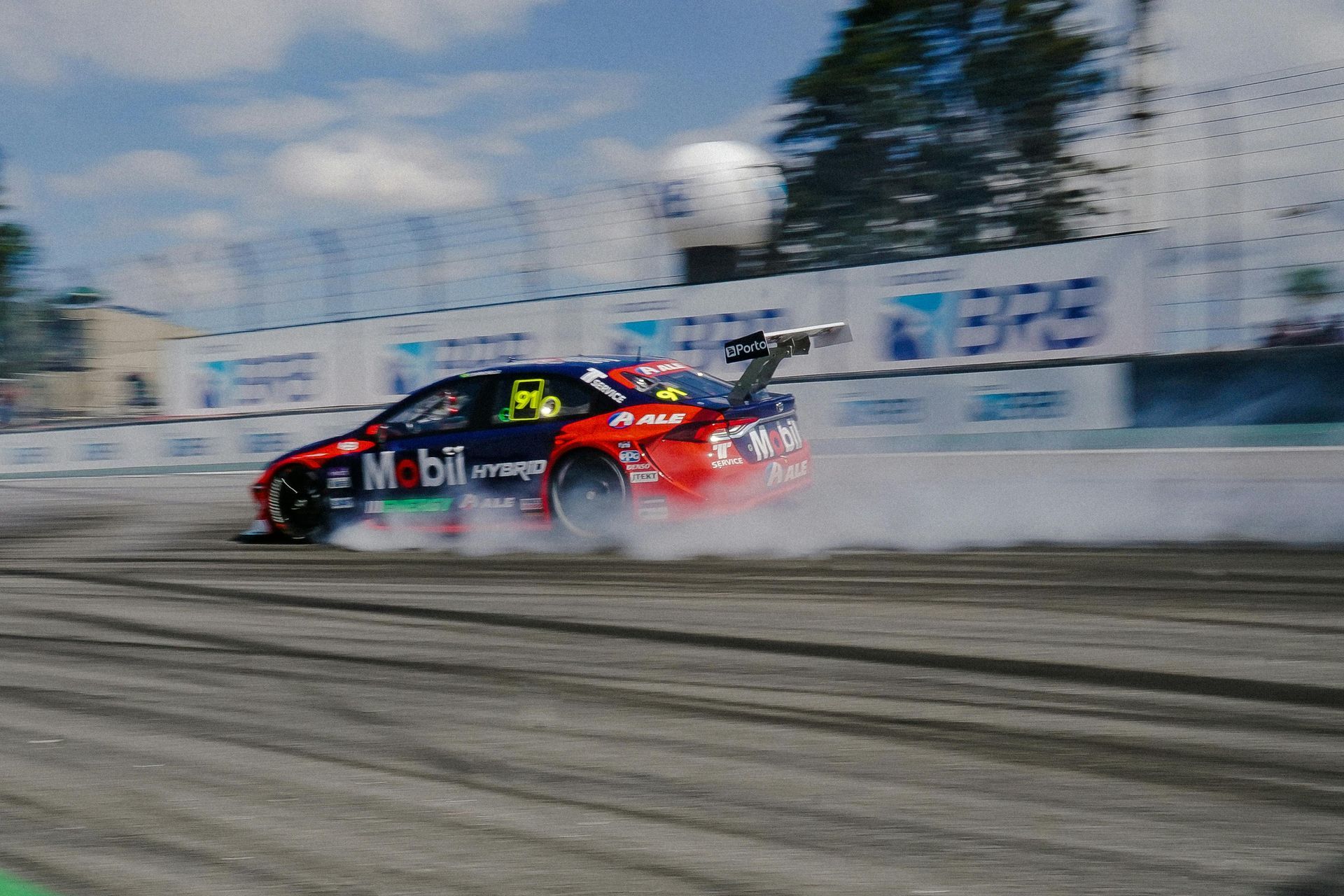 A red and blue Mobil race car drifts around a track curve, kicking up white smoke against a backdrop of BRB signs.