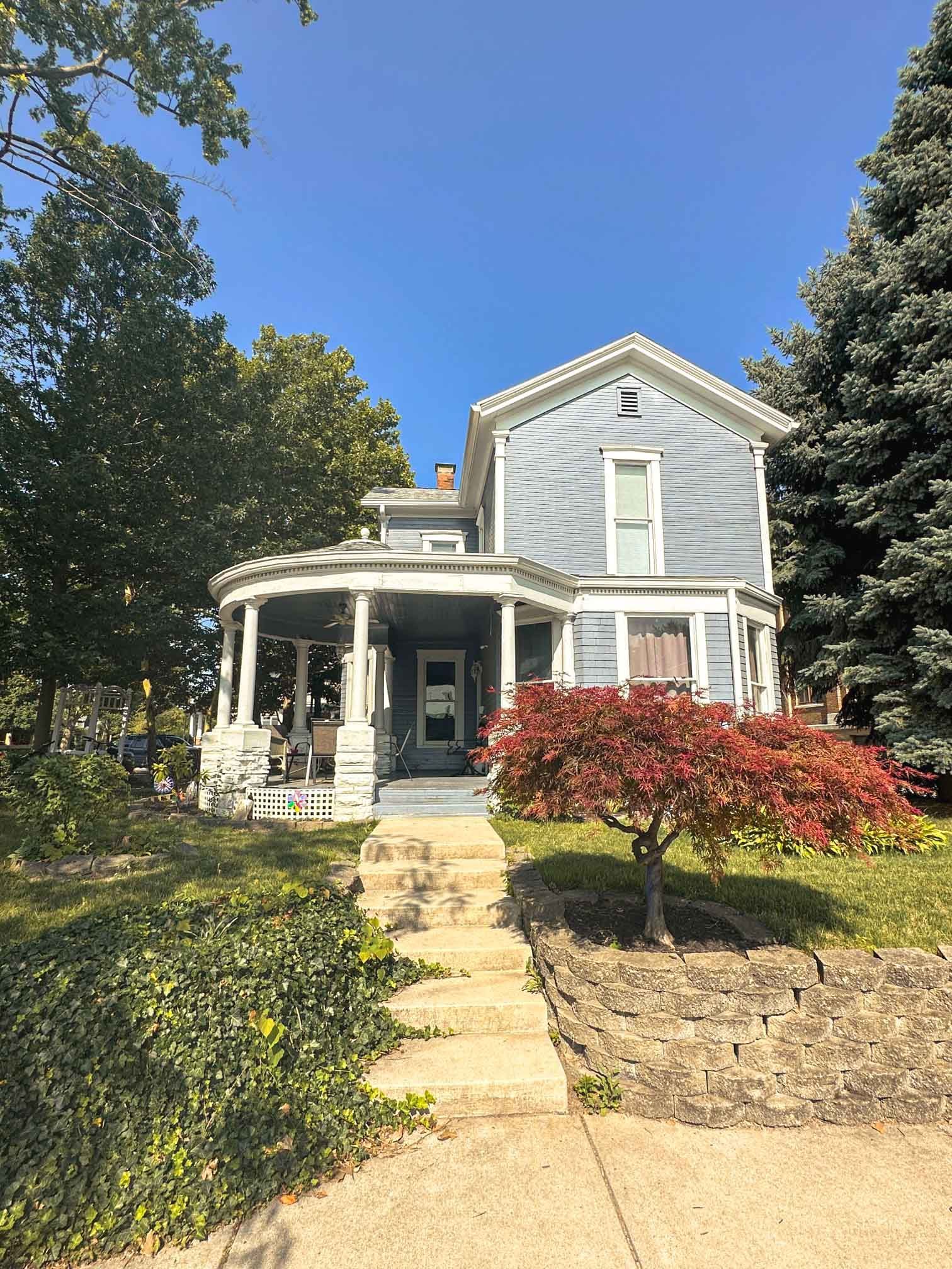 A large house with a porch and stairs is surrounded by trees on a sunny day.
