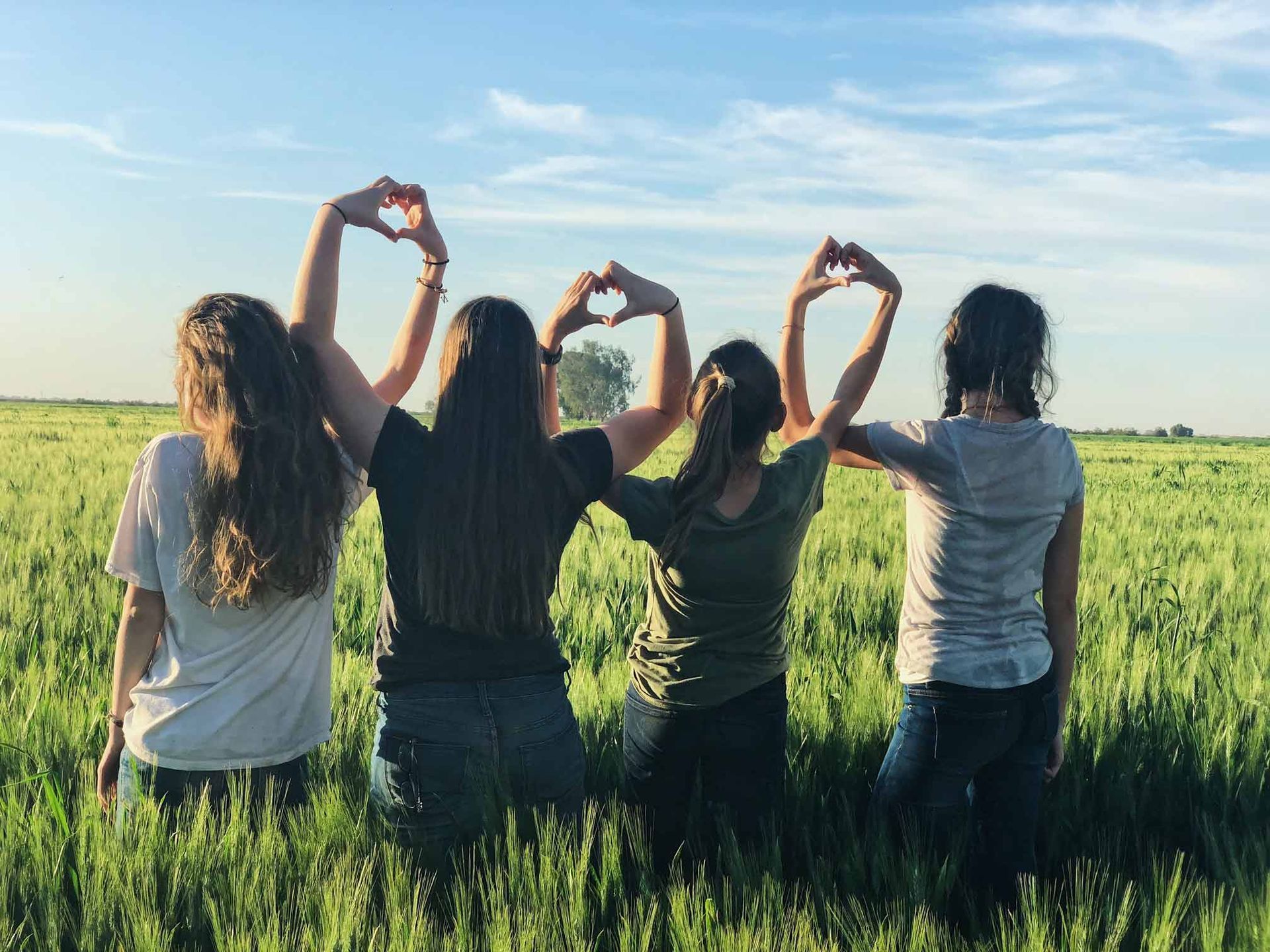 Four women are making a heart shape with their hands in a field.