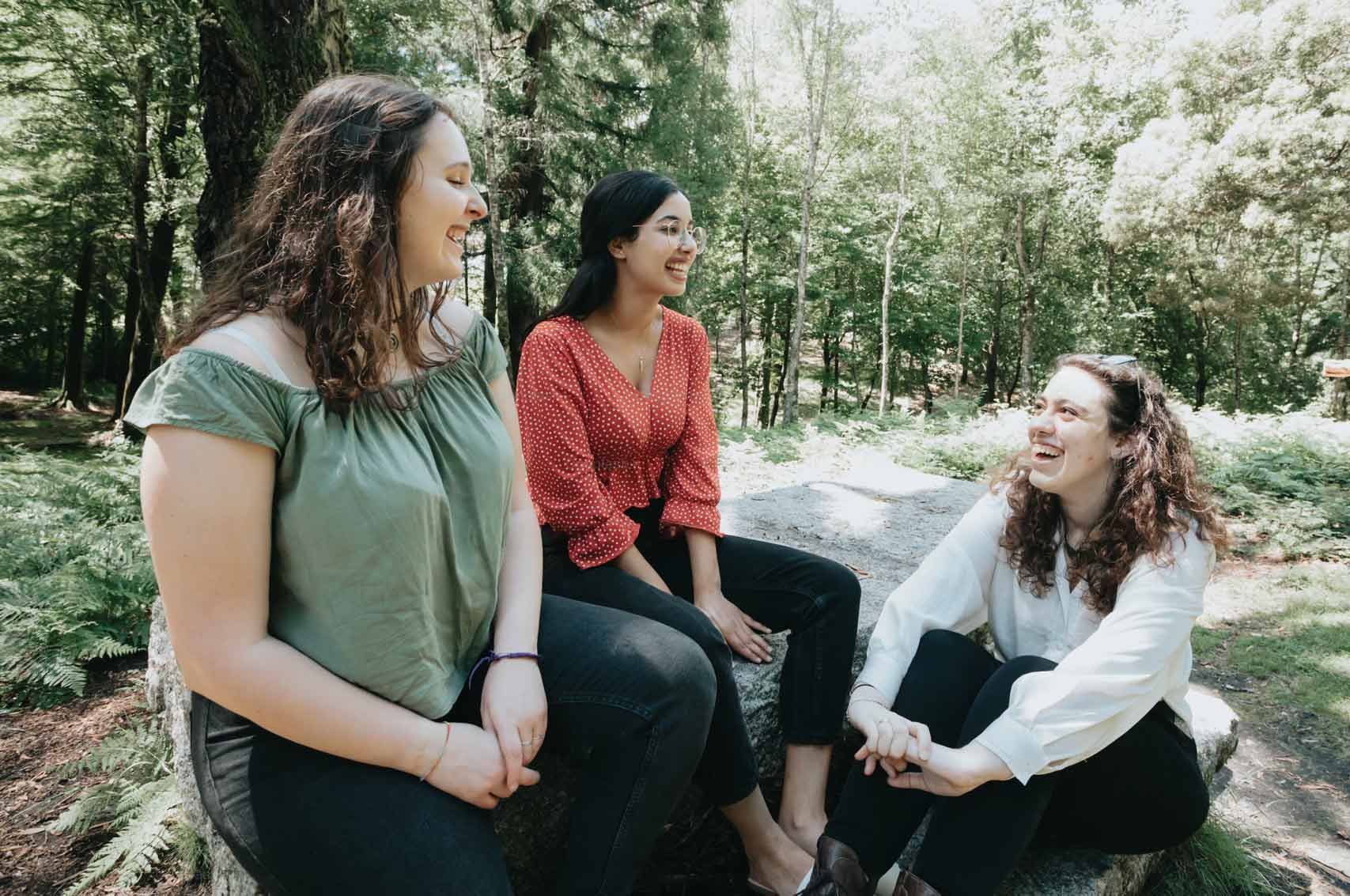 Three women are sitting on a rock in the woods talking to each other.