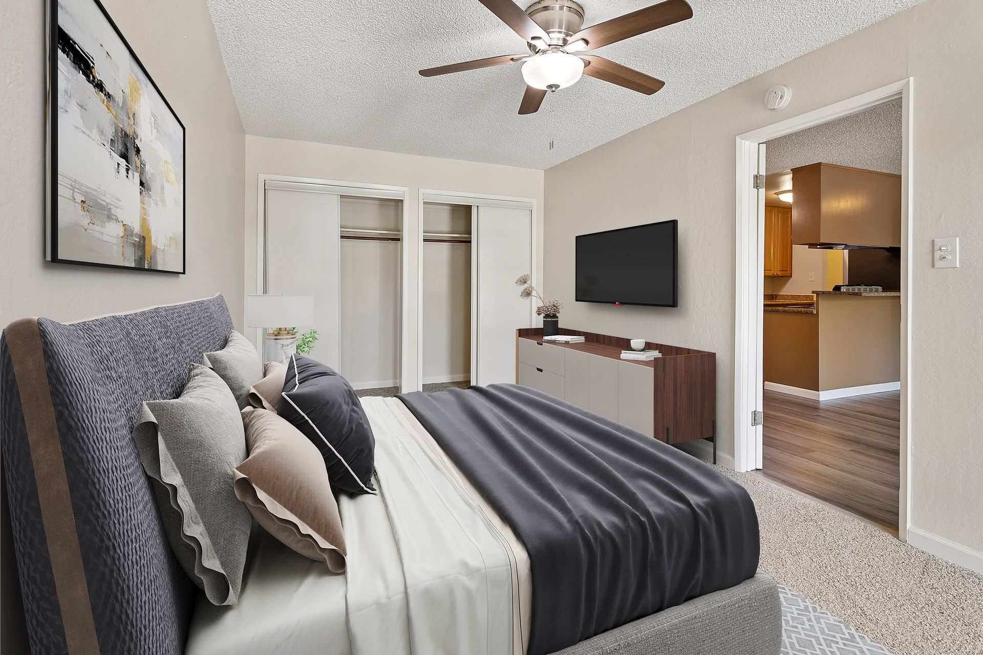 A furnished bedroom with a gray bed, ceiling fan, television, and an open doorway leading to a kitchen at Parkview Apartments.