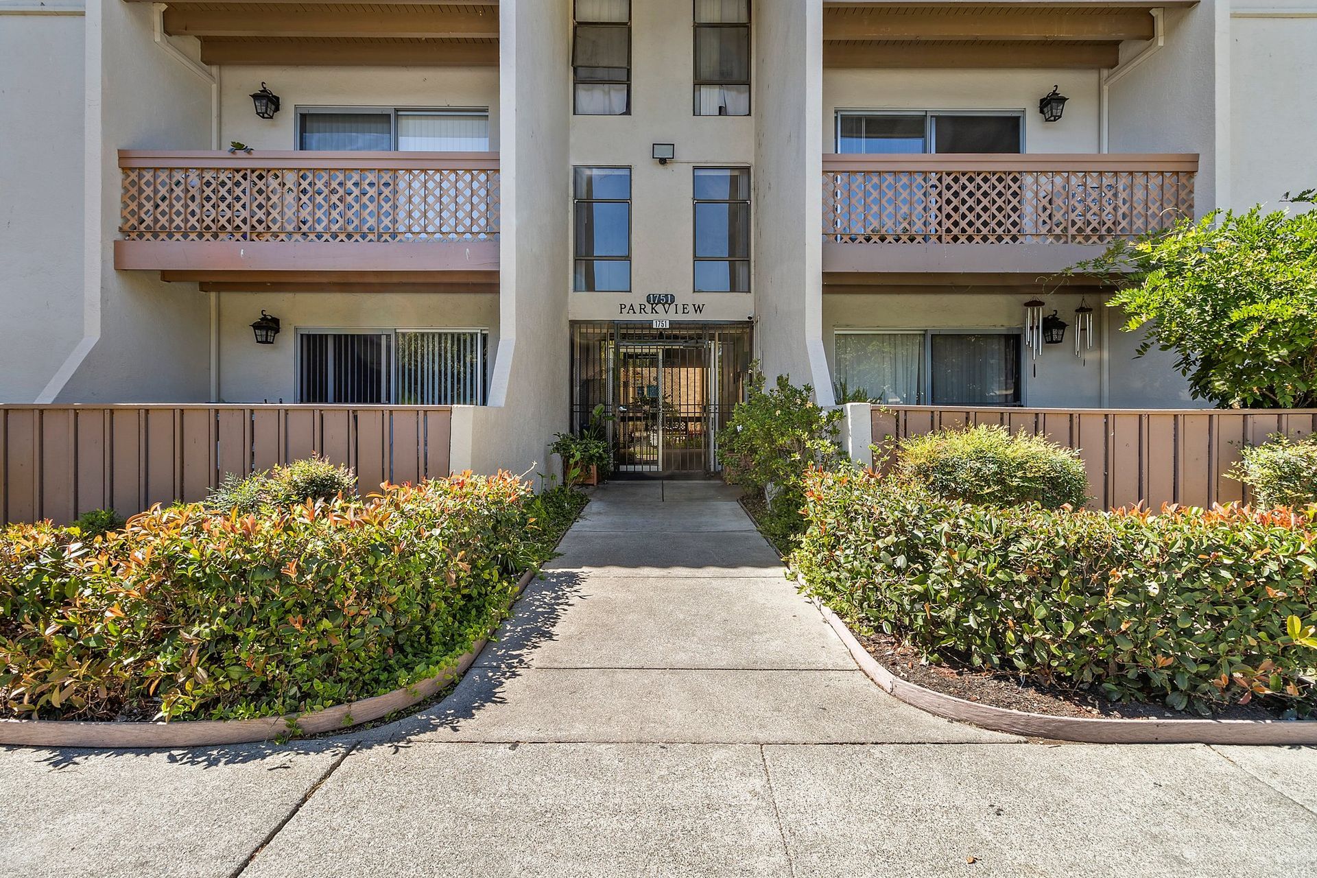 A lush, tree-lined park surrounds a calm pond under a blue sky, with a road and parked cars along the bottom edge at Parkview Apartments.