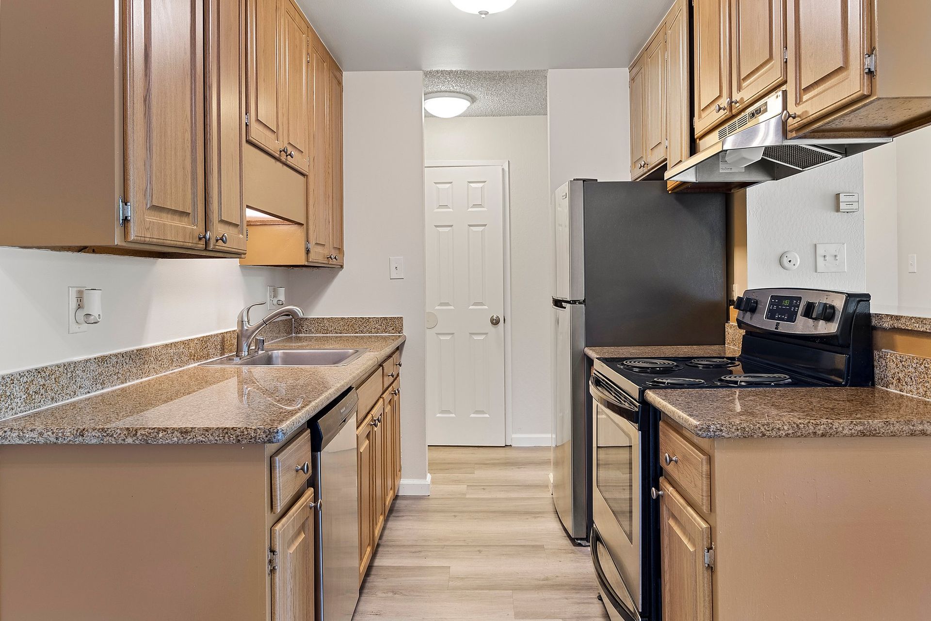 A galley-style kitchen with light wood cabinets, granite countertops, stainless steel appliances, and wood-look flooring  at Parkview Apartments.