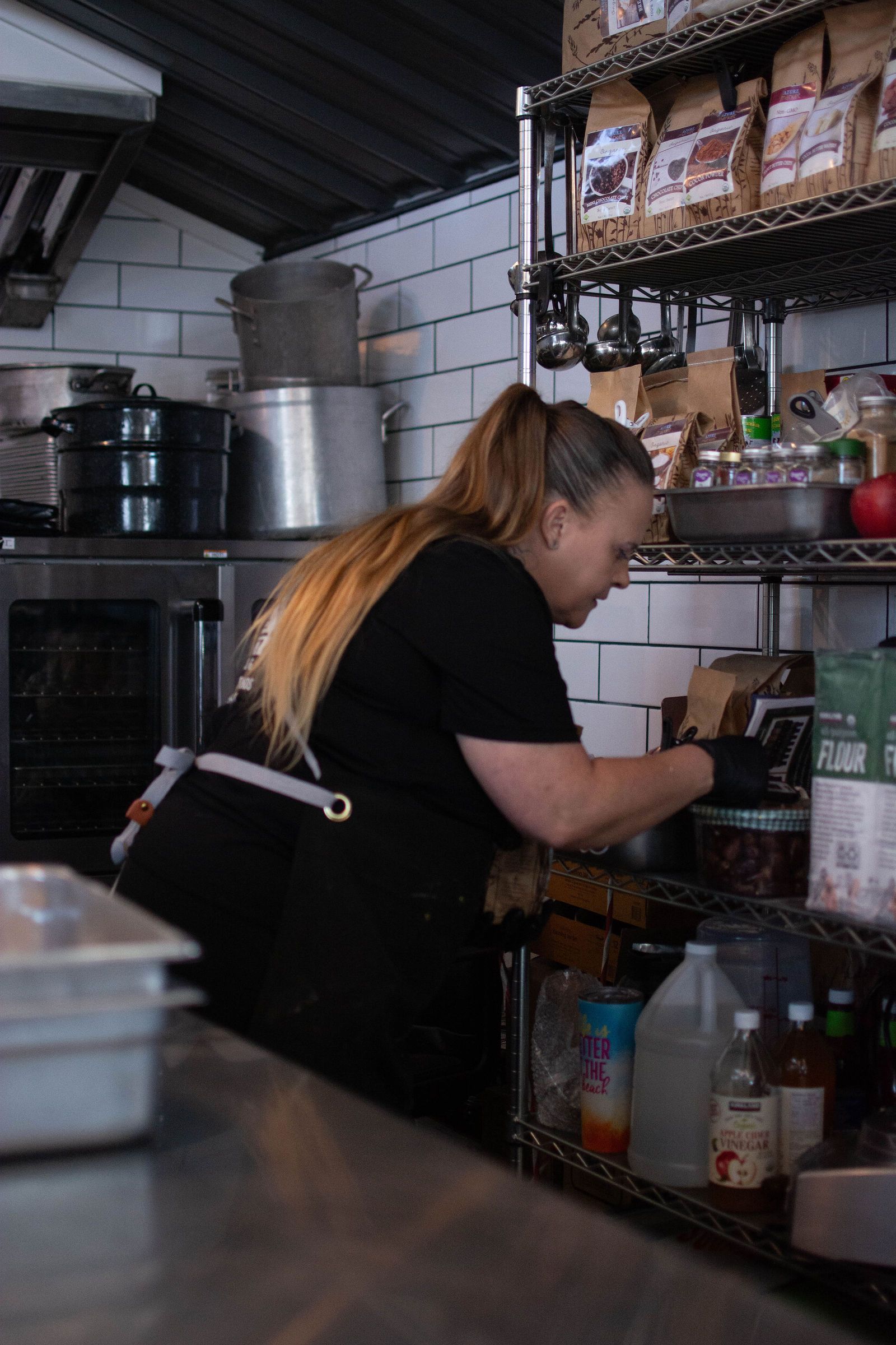 Woman in black apron at a commercial kitchen, reaching for a product on shelf.