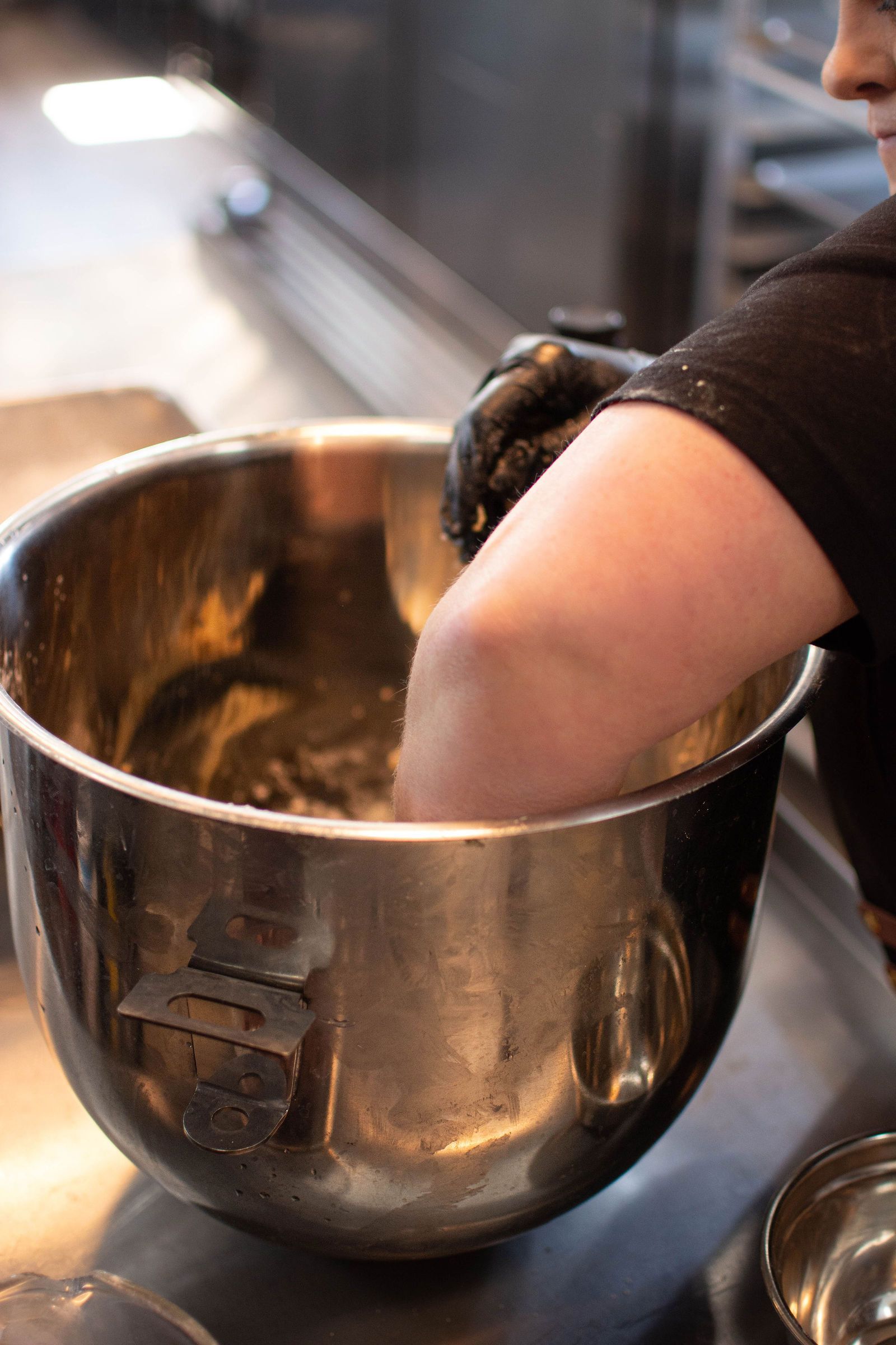 Person in black glove stirring batter in a large metal bowl in a kitchen.