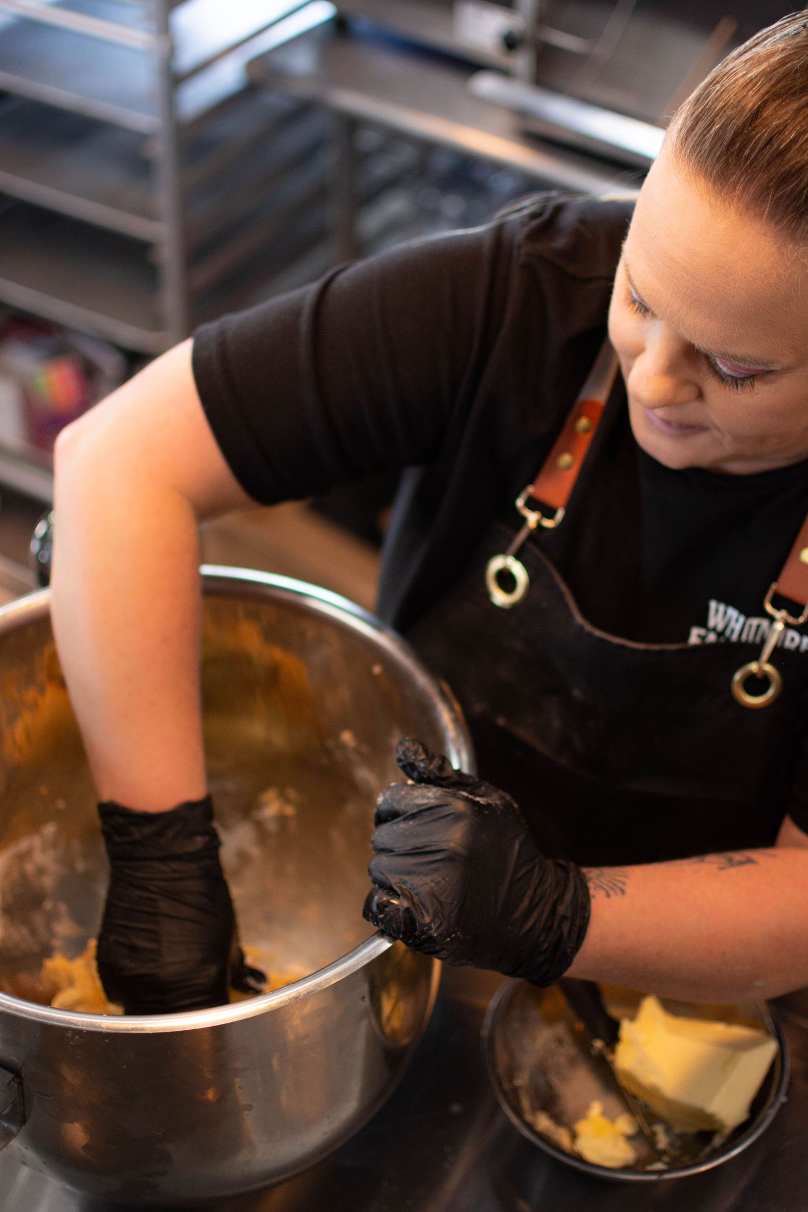Person wearing black gloves mixing dough in a metal bowl, wearing a black apron in a kitchen.