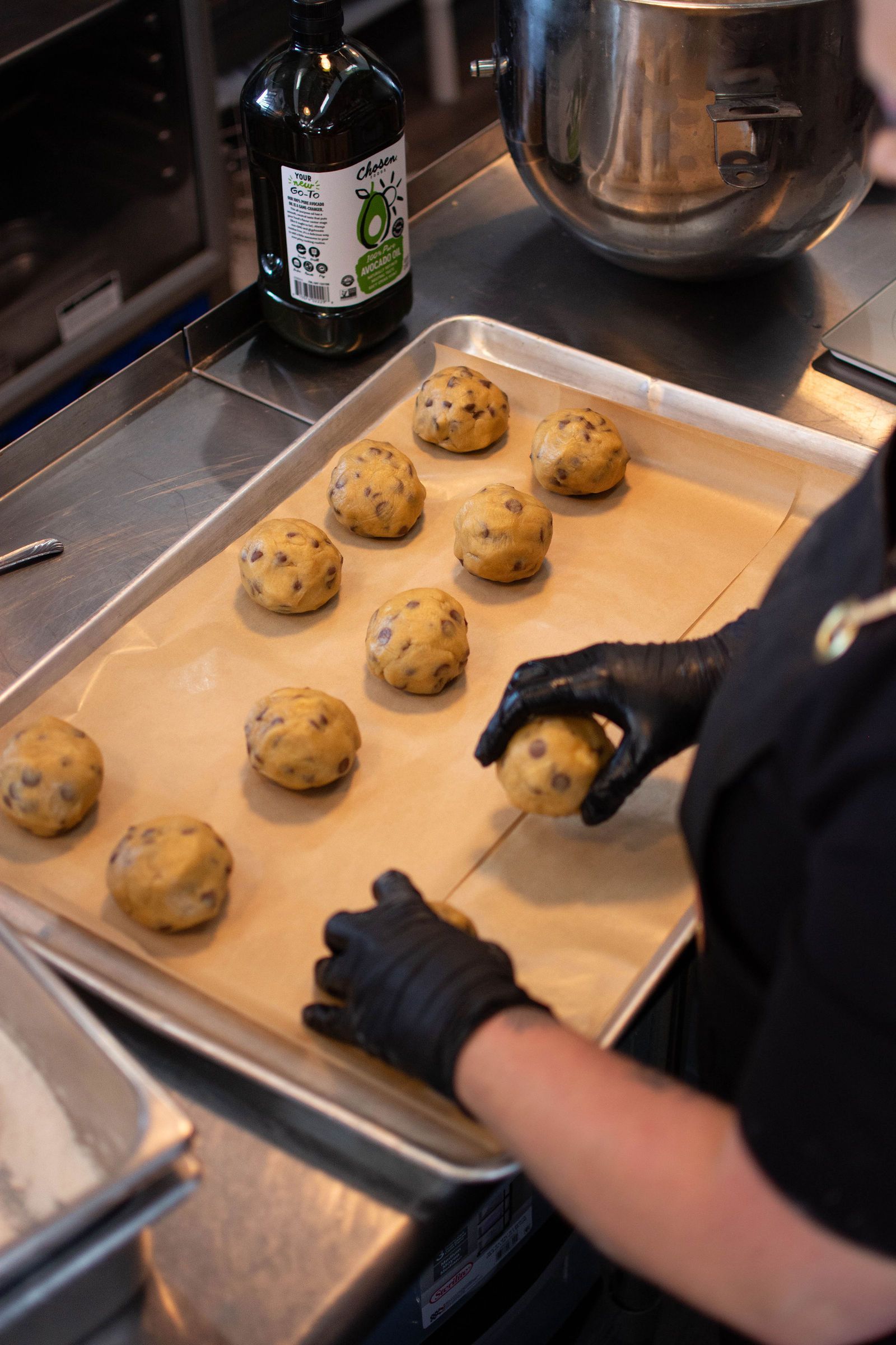 Hands in black gloves place cookie dough balls on a baking sheet lined with parchment paper.