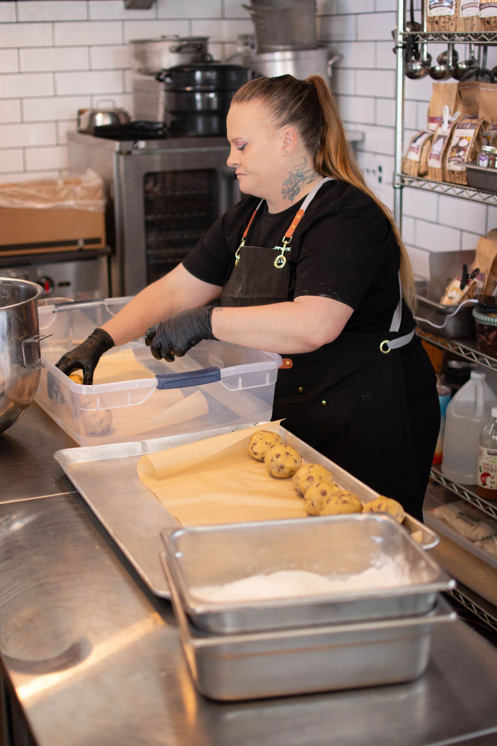 Woman in black shirt and apron prepares cookies in a commercial kitchen, using containers and baking sheets.