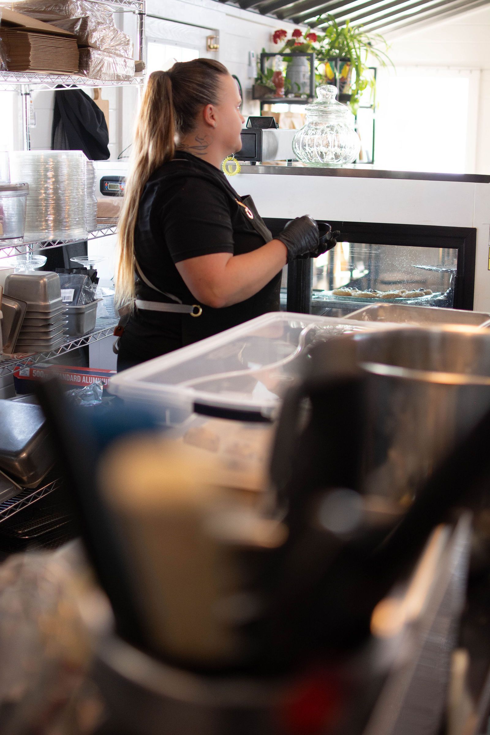 Woman in black shirt and gloves, ponytail, in restaurant kitchen, near serving area.